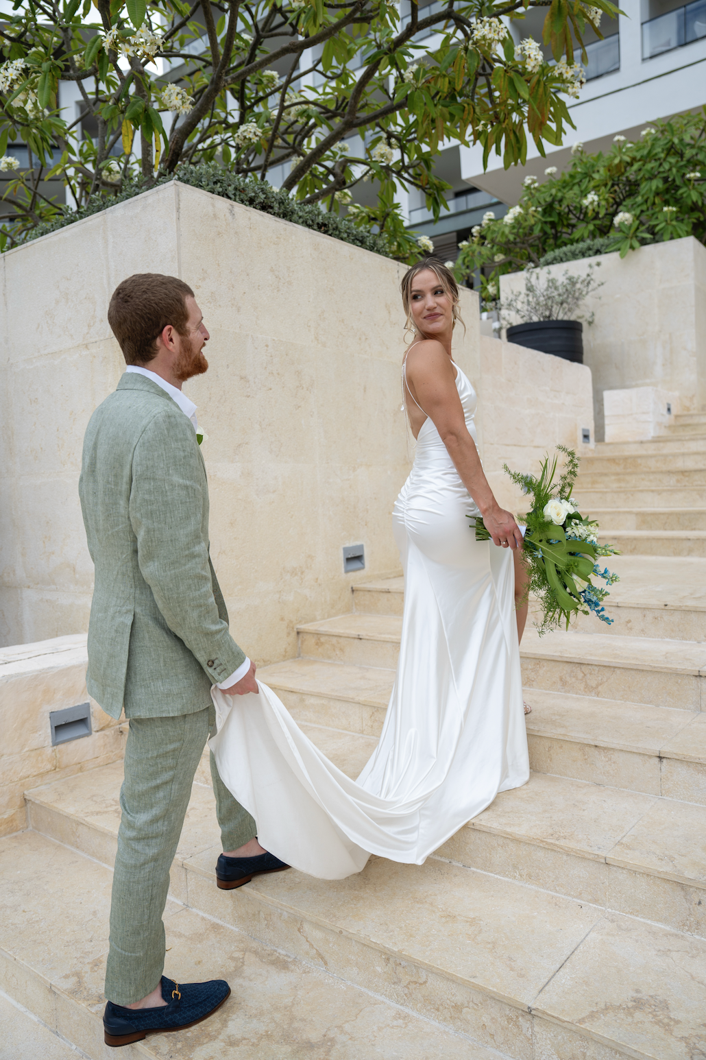 A bride with a bouquet of flowers stands on stairs, looking back at her groom who is holding her dress.