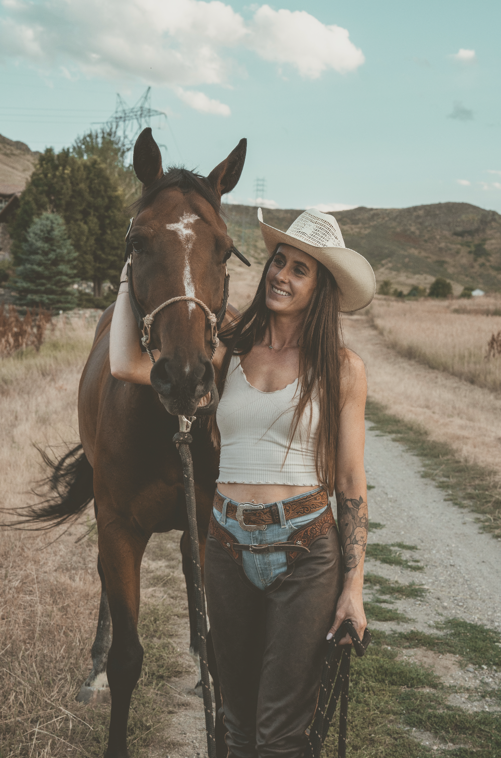 A woman smiling and wearing a cowboy hat stands next to a brown horse on a dirt path in a rural landscape with mountains and a partly cloudy sky.