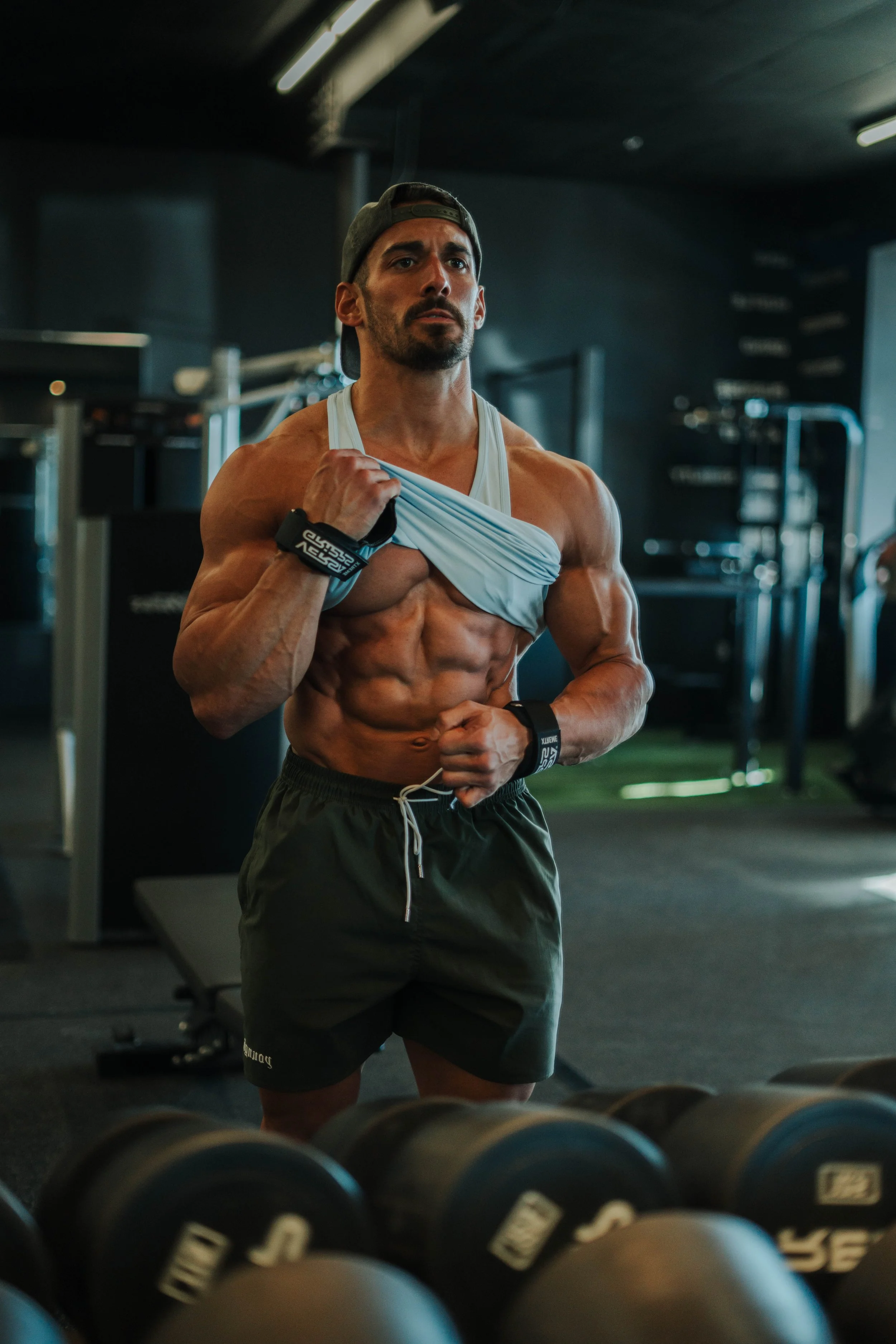 Muscular man in a gym lifting his shirt and showing off his abs, wearing a backwards cap, shorts, and wristbands.