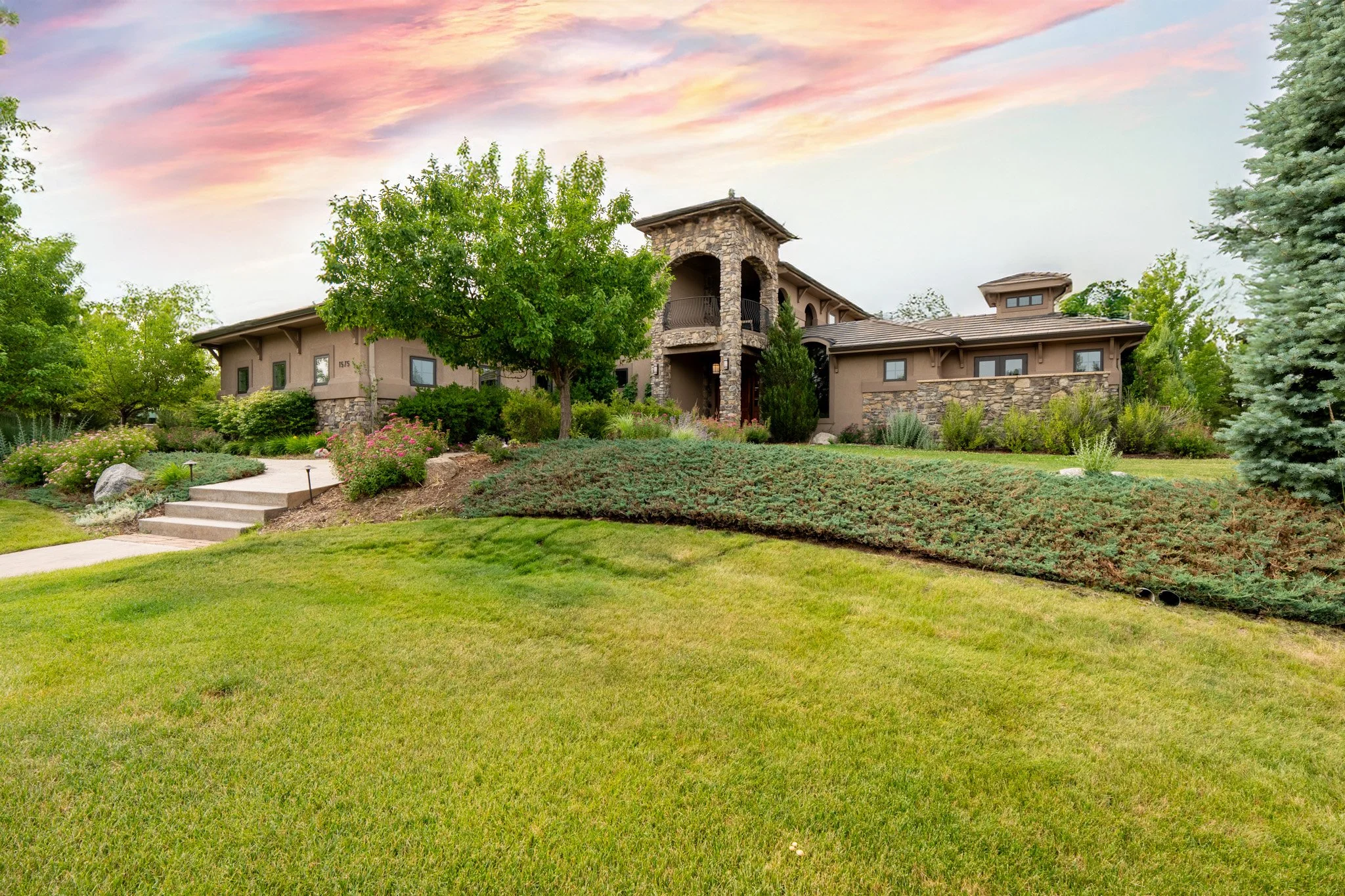 A large, modern house with stone accents, surrounded by green landscaping, trees, and a well-maintained lawn under a colorful sky.
