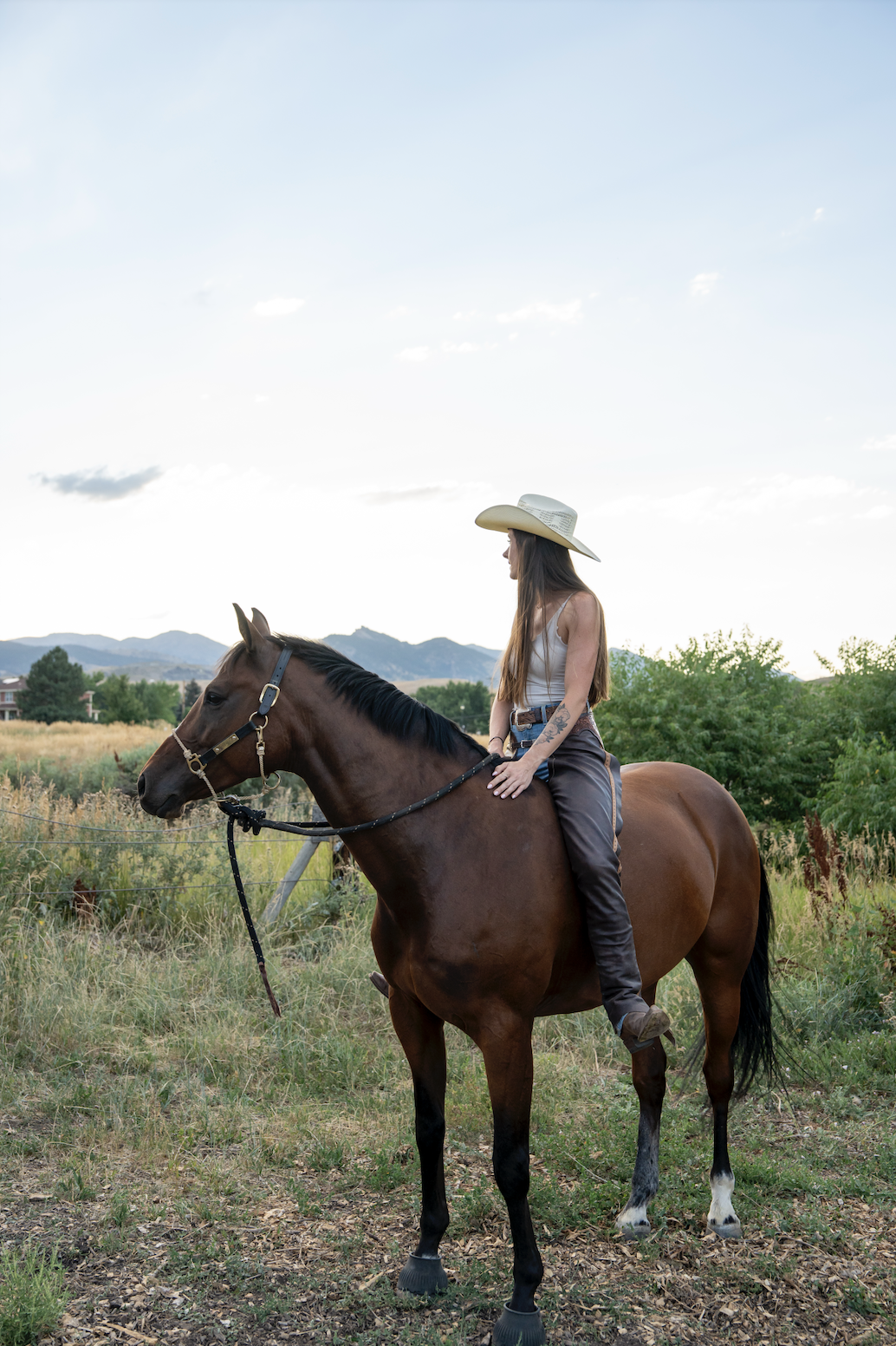 A woman wearing a cowboy hat, sleeveless top, and jeans sits on a brown horse in an open field with mountains and trees in the background.