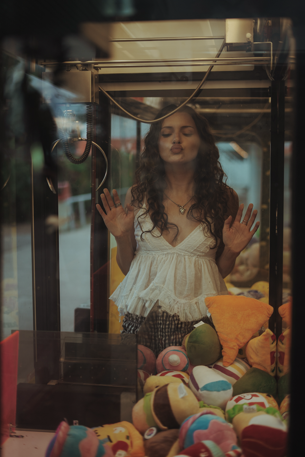 A woman with curly hair and wearing a white sleeveless top is pressing her hands against a glass enclosure in a claw machine, appearing to try and grab plush toys inside.