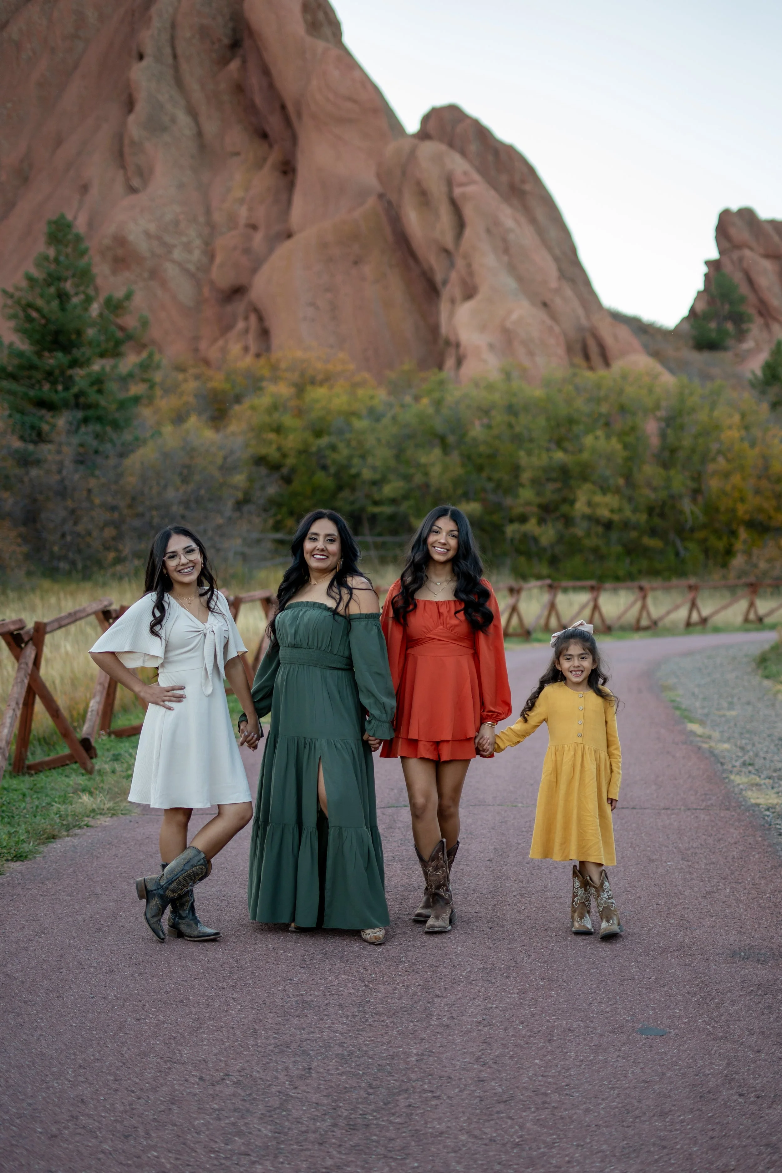 Group of four women and a girl holding hands, walking outdoors on a path with rocky mountains and trees in the background. Family photos