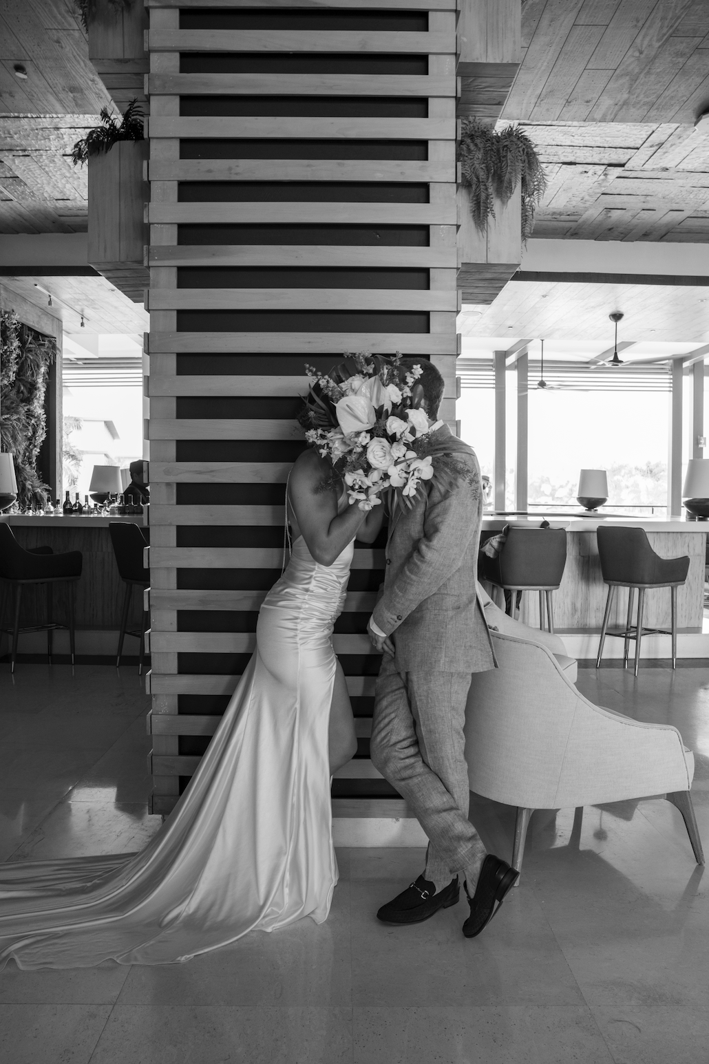 A black and white photo of a bride and groom sharing a romantic moment behind a wooden partition; the bride is in a satin wedding gown, and the groom is dressed in a tuxedo.