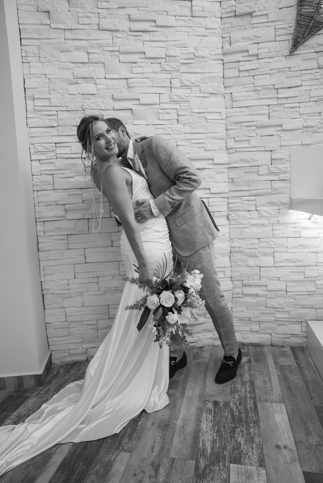 A bride and groom embracing and smiling during their wedding, with the bride holding a bouquet of flowers, standing against a textured white brick wall.