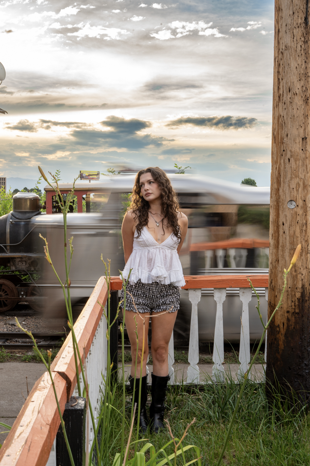 A young woman with curly hair standing outdoors near a wooden pole and a white railing, with a moving train in the background during sunset.