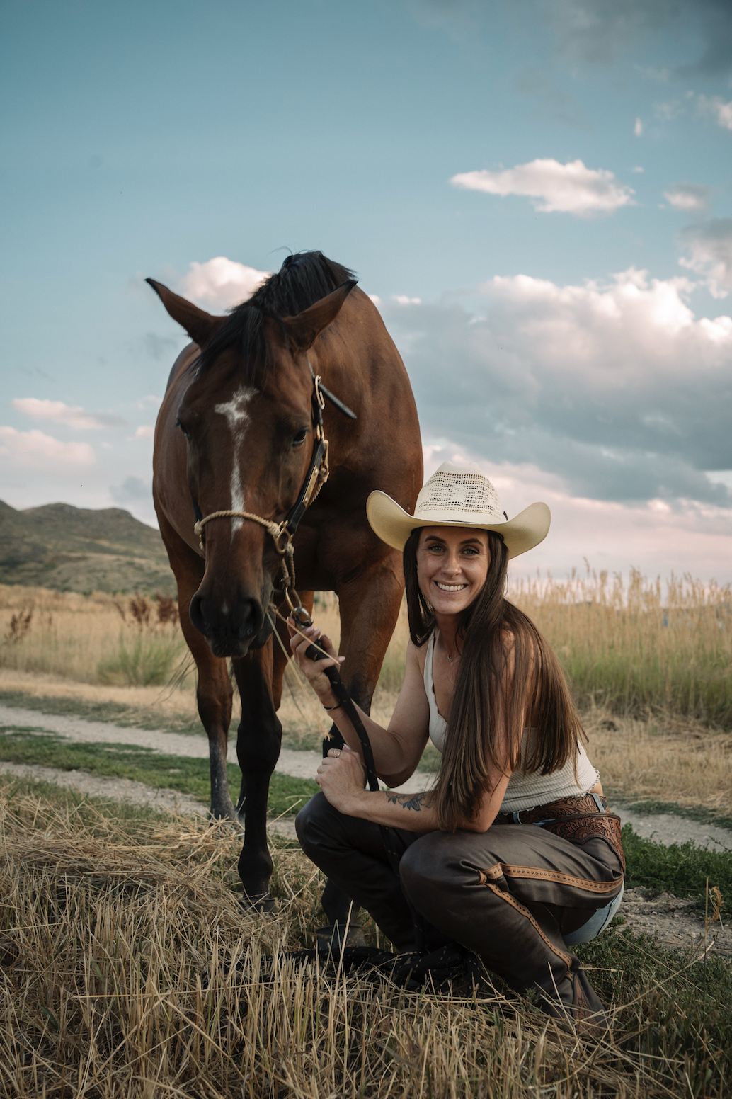 A woman with long brown hair, wearing a white cowboy hat, squatting next to a brown horse, holding its reins and smiling outdoors in a grassy field with blue sky and clouds.