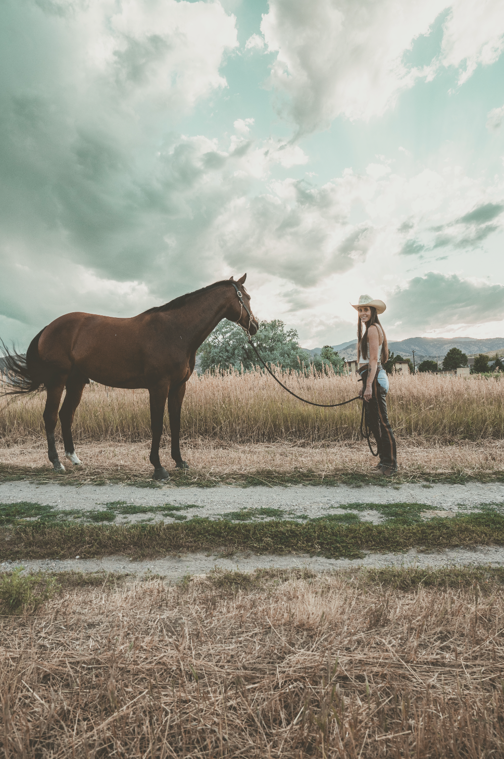 A woman wearing a cowboy hat, standing in a field holding a horse's reins. The sky is cloudy with mountains in the background.