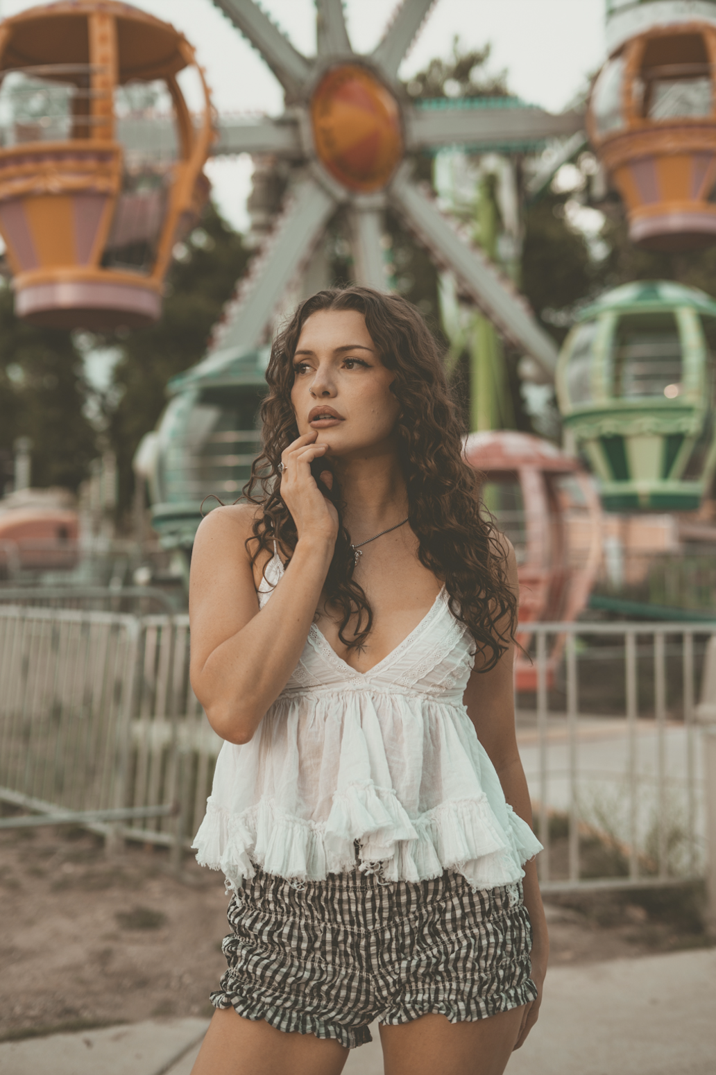 A woman with curly brown hair, wearing a white sleeveless top and checkered shorts, stands in front of a colorful amusement park Ferris wheel, looking thoughtfully to the side.