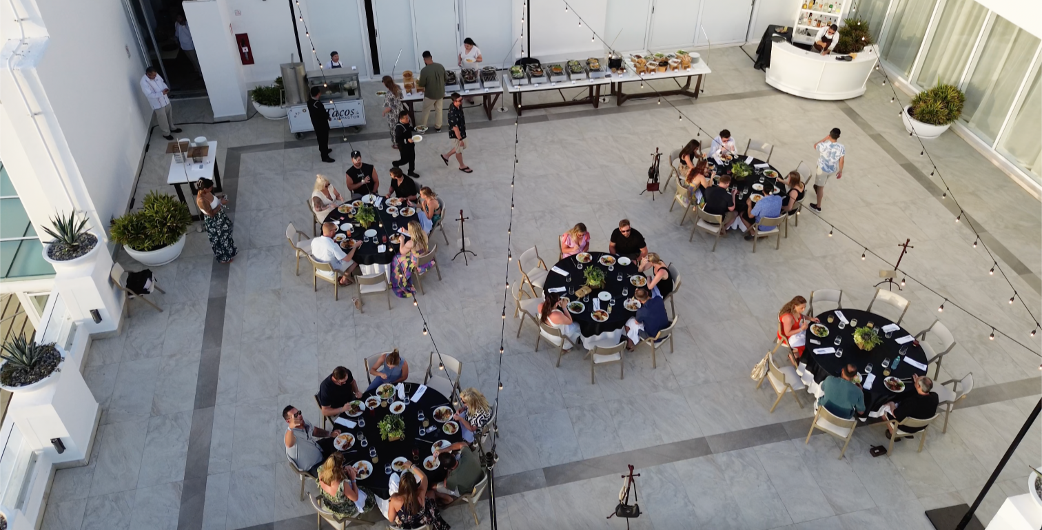 People dining at round tables with black tablecloths on a spacious indoor terrace, with a buffet of food along the back wall, potted plants, and string lights hanging above.