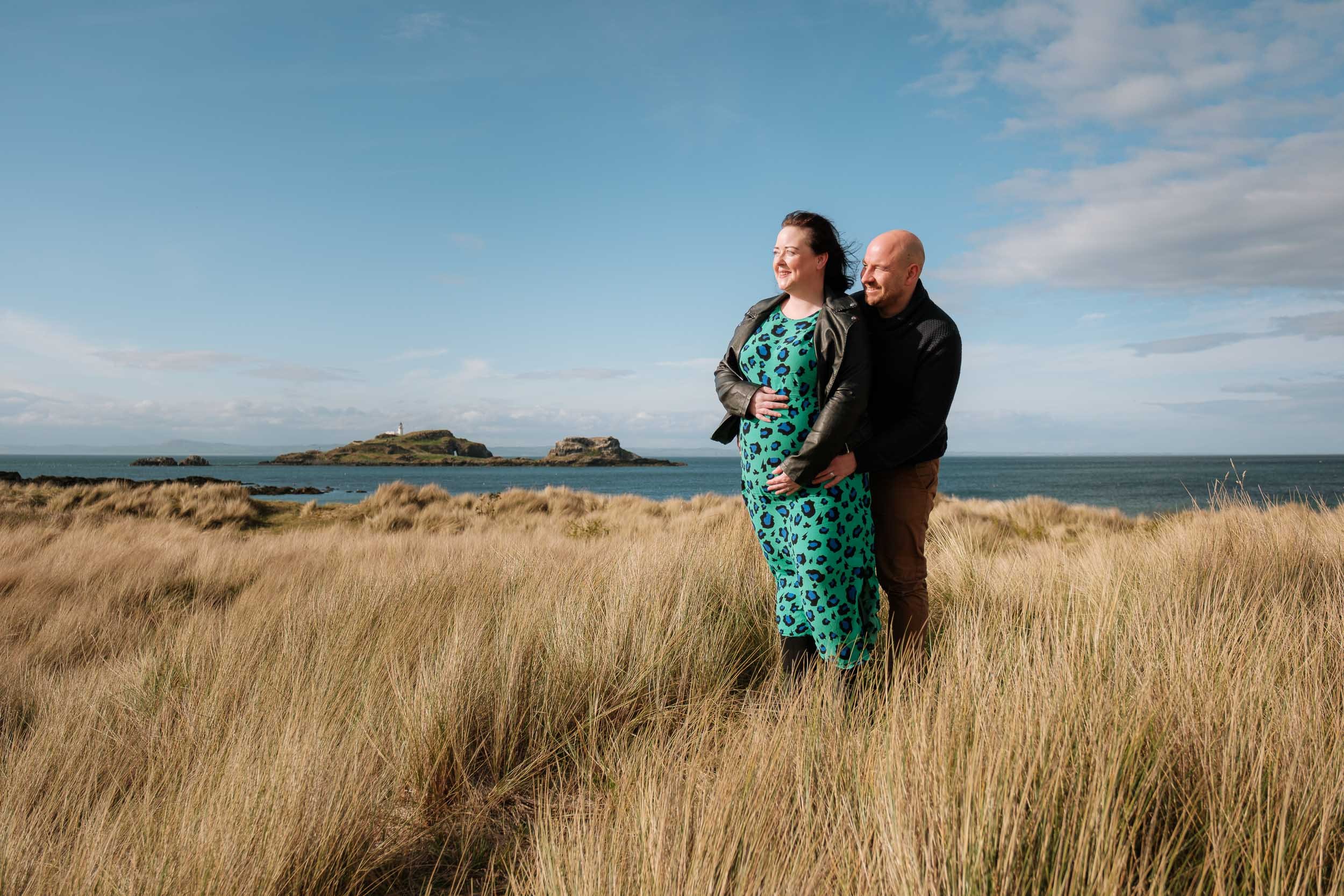 A couple standing in a grassy field near the ocean, with an island and lighthouse in the distance, under a partly cloudy sky.