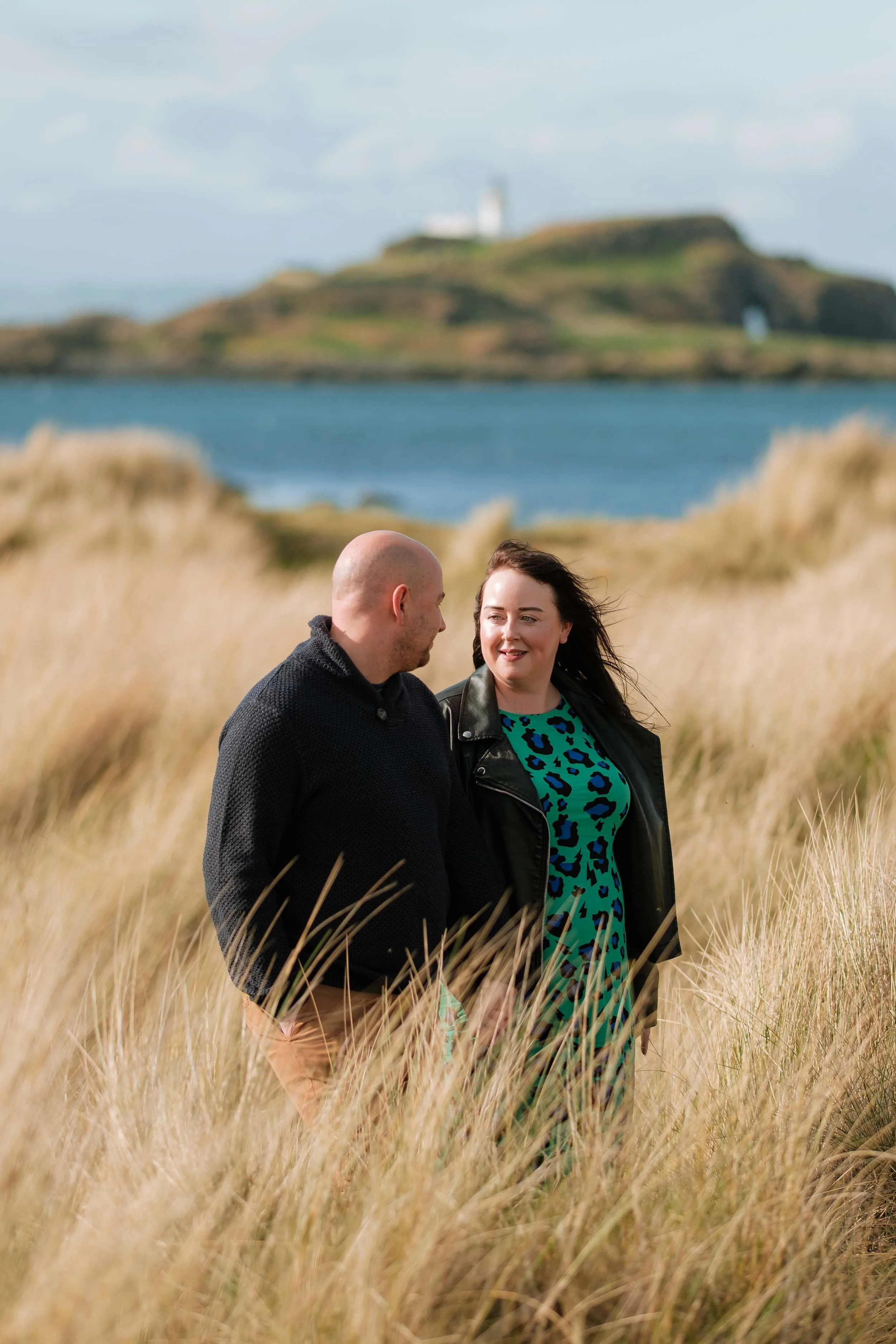 A couple walking through tall golden grass near a body of water with a hill and a lighthouse in the background.