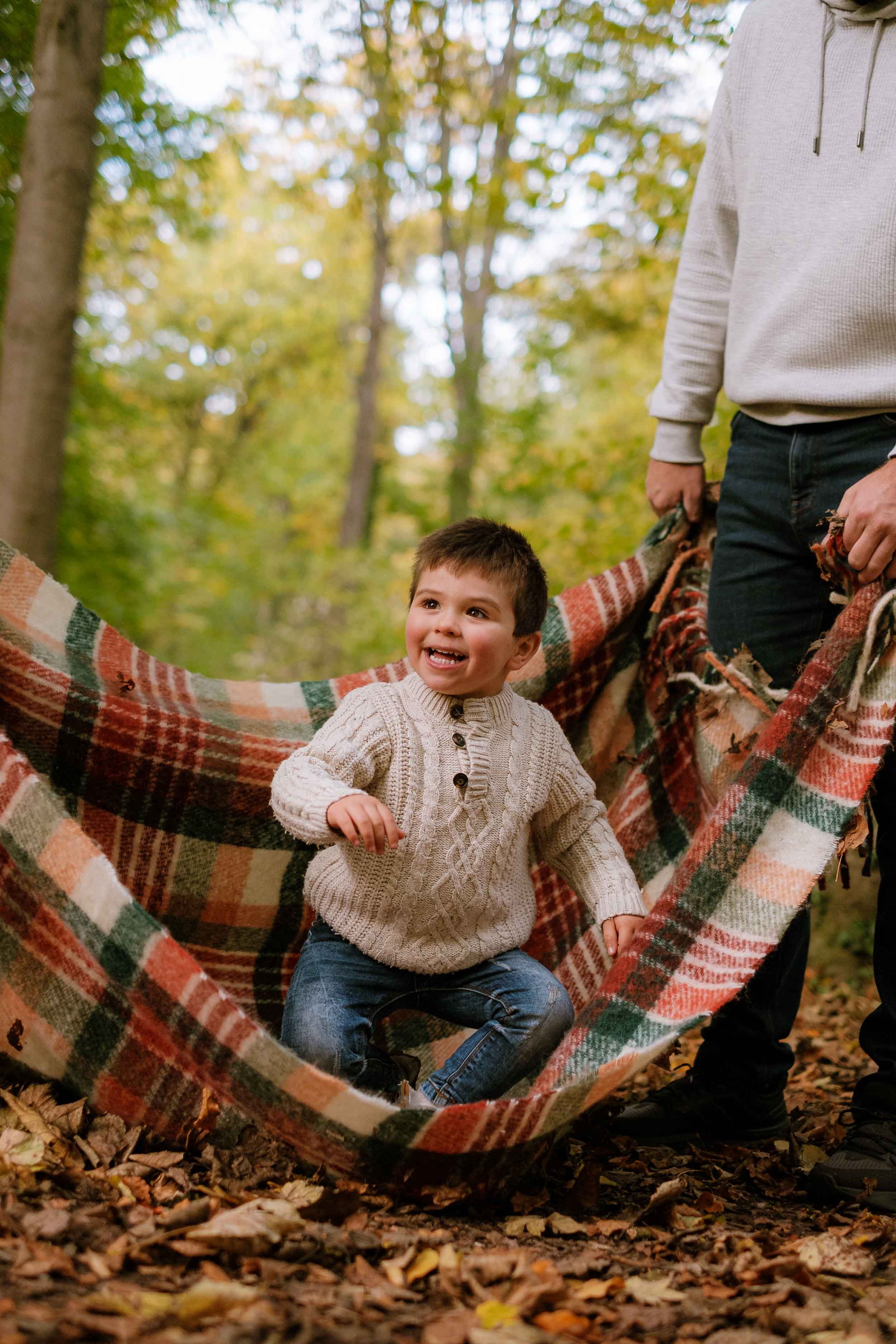 A young boy in a cream cable-knit sweater and jeans sitting in a plaid hammock on a fall day, surrounded by trees with autumn leaves, holding hands with a person standing nearby.