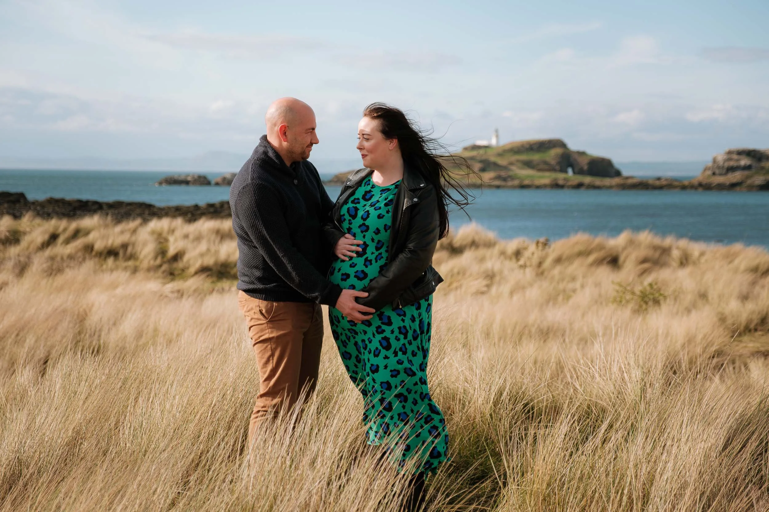 A couple stands in sand dunes near the coastline of East Lothian, with the man gently holding the woman’s pregnant belly during a maternity photoshoot.  In the background, there is a body of water, rocky islands, and a lighthouse.