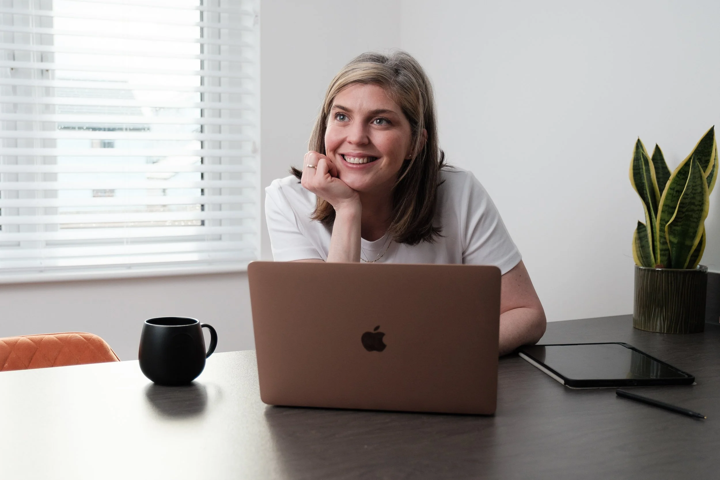 A woman smiling while sitting at a desk with an open MacBook, a black mug, a tablet, a pen, and a potted plant in a bright room.
