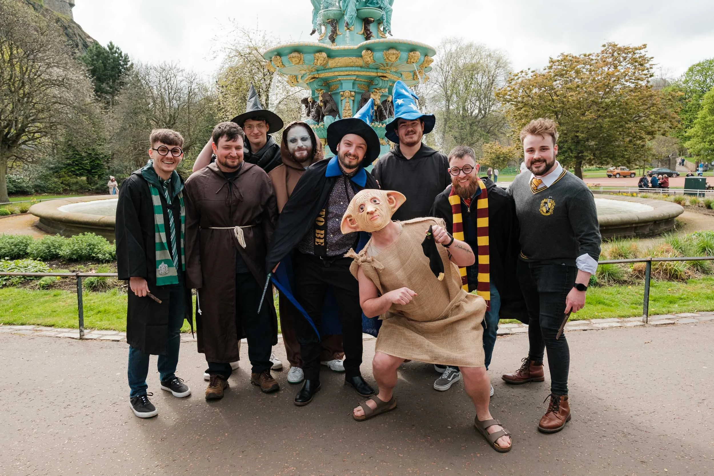 Group of people dressed as witches and Harry Potter characters posing near a decorative fountain in a park.