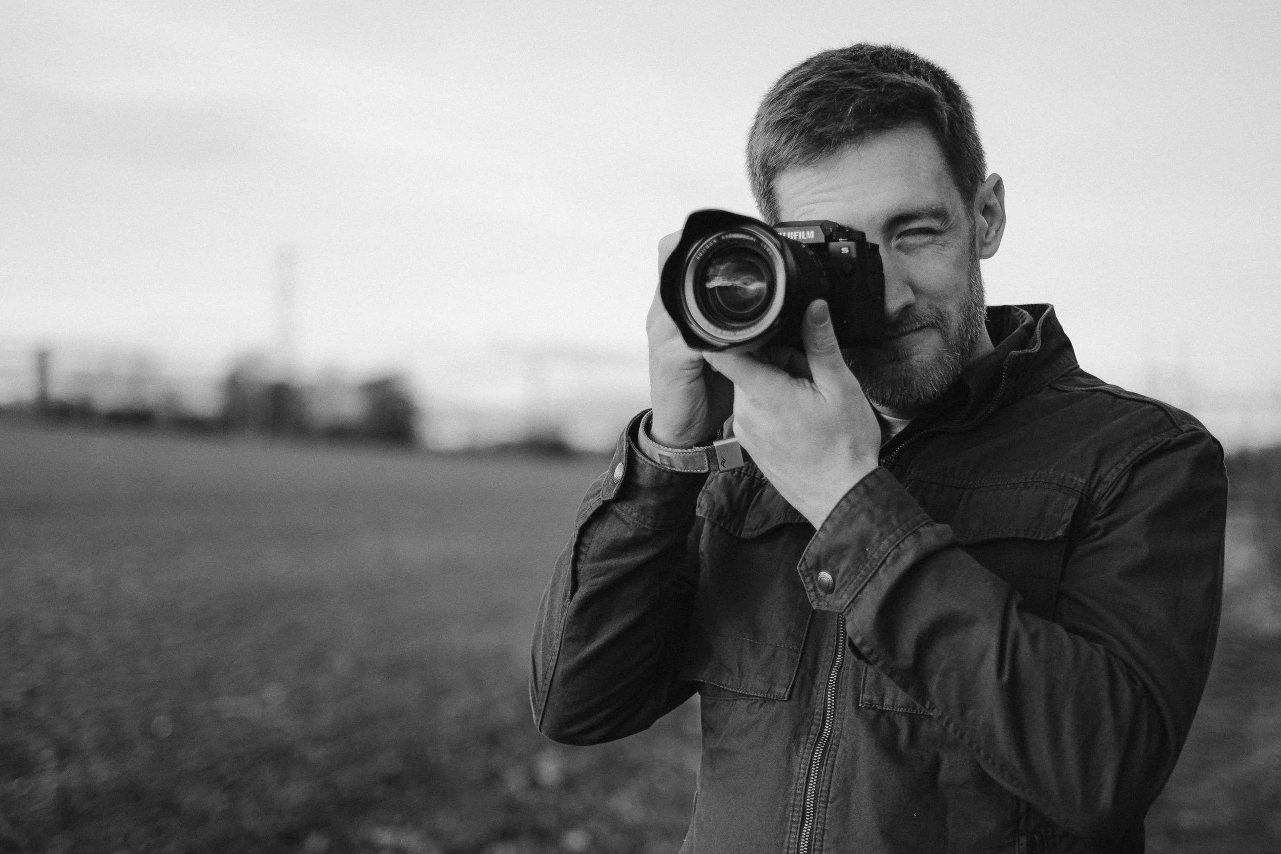A man outdoors in Midlothian taking a photograph with a camera, wearing a jacket, with a blurred background.