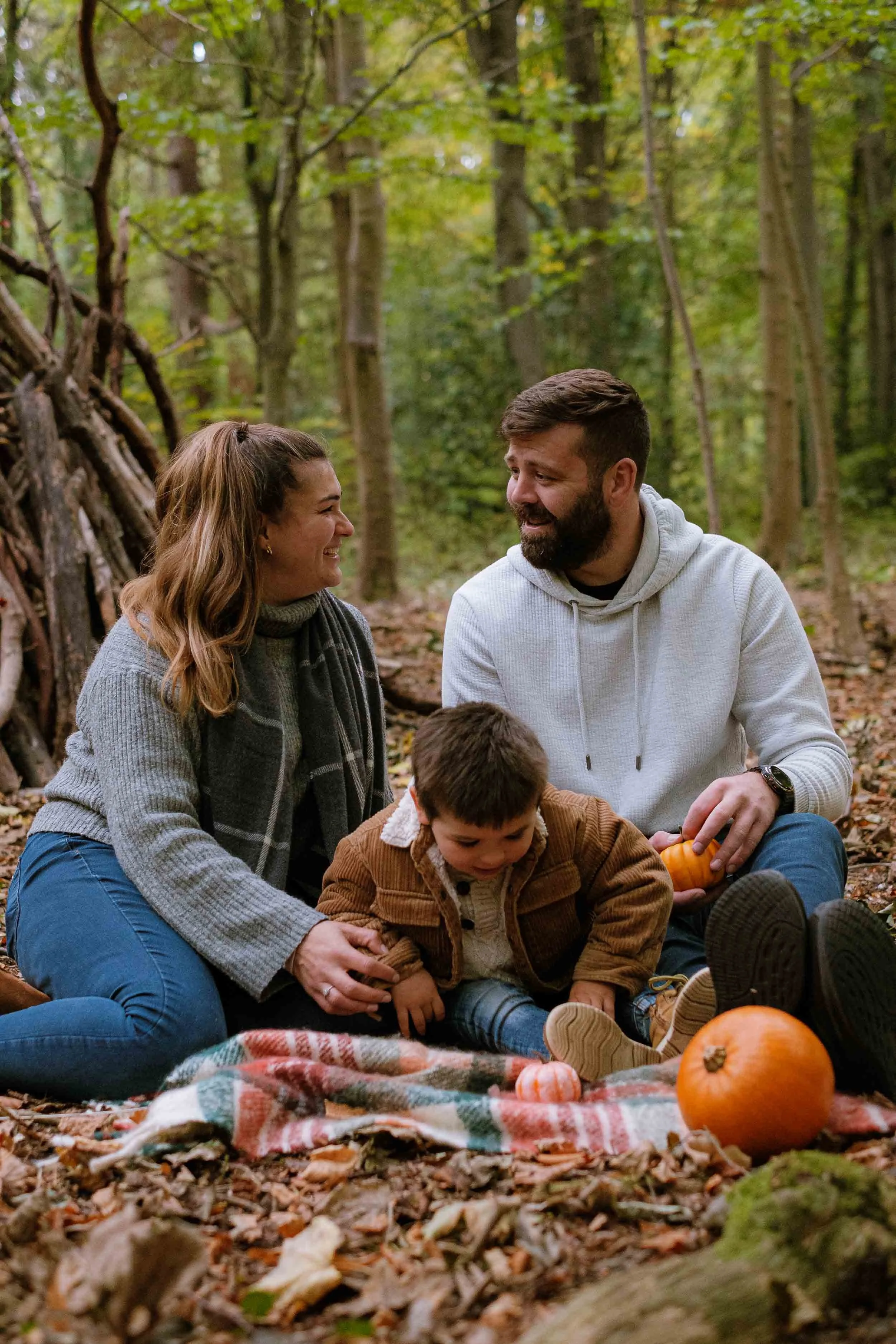 A photograph of a family sitting on a blanket in the woods during autumn in Midlothian, surrounded by pumpkins.