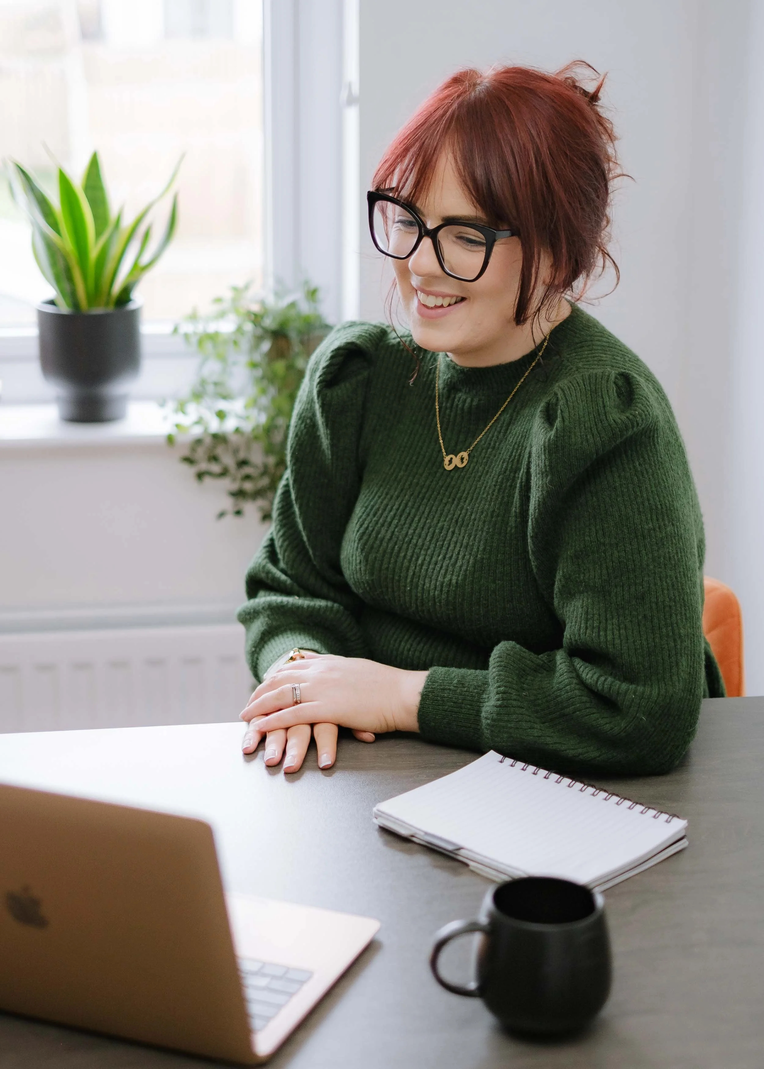 A female business owner portrait during personal brand photography session in Edinburgh, sitting at a table near a window, smiling and looking at her laptop, with a notebook and a black coffee mug on the table, and plants in the background.