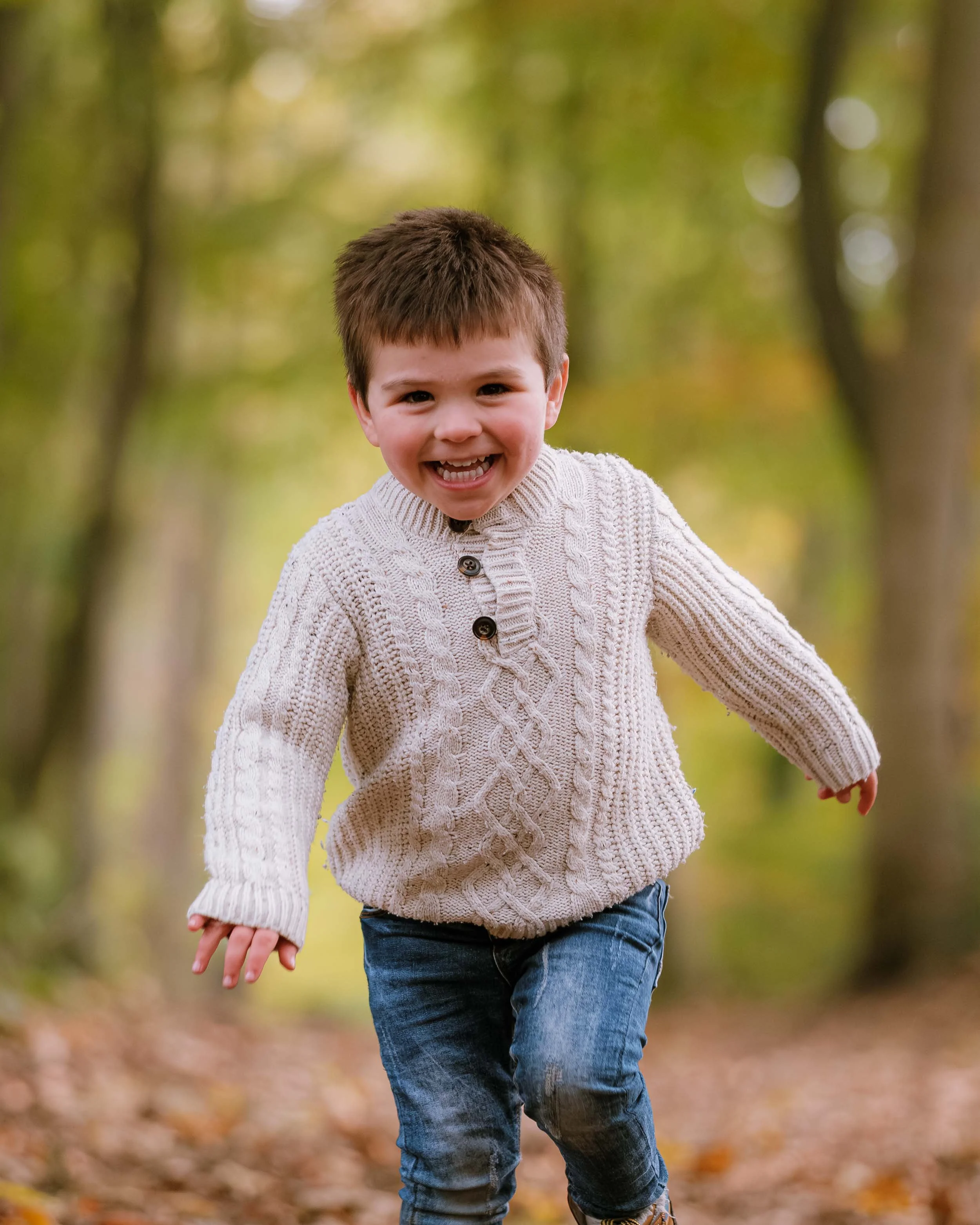 Young boy smiling and running in a forest during fall, wearing a cream-colored cable knit sweater and jeans.