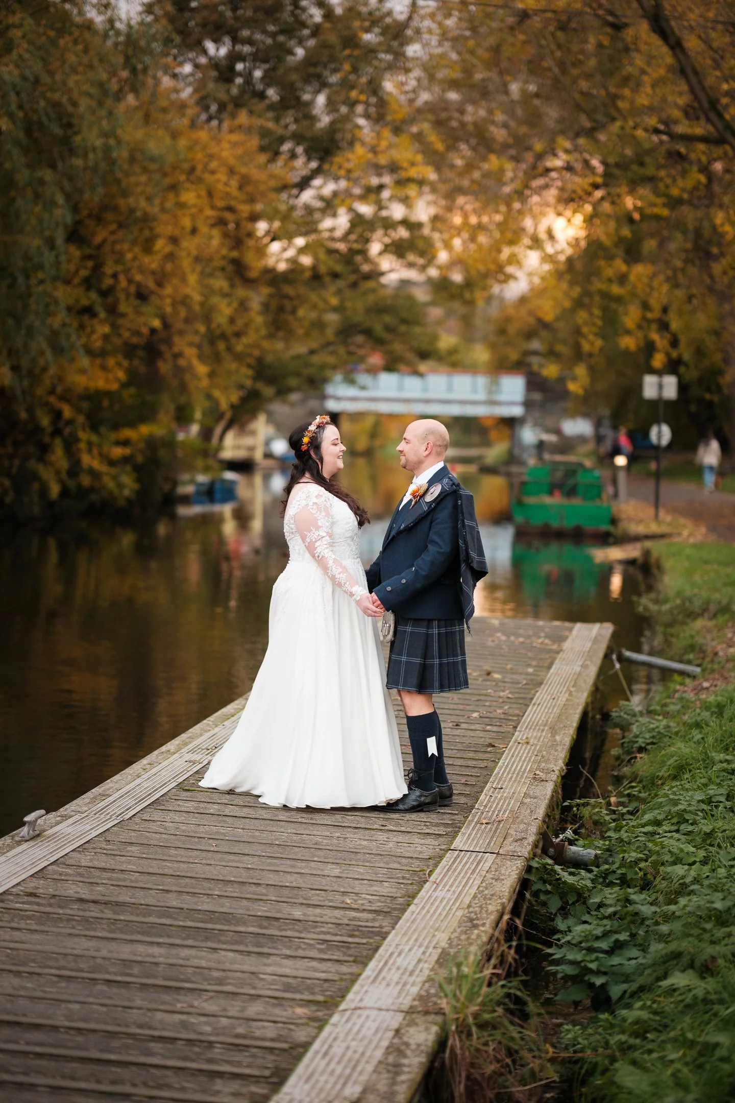 A bride and groom standing on a wooden dock by a river in Edinburgh, holding hands and smiling at each other during their wedding photos.