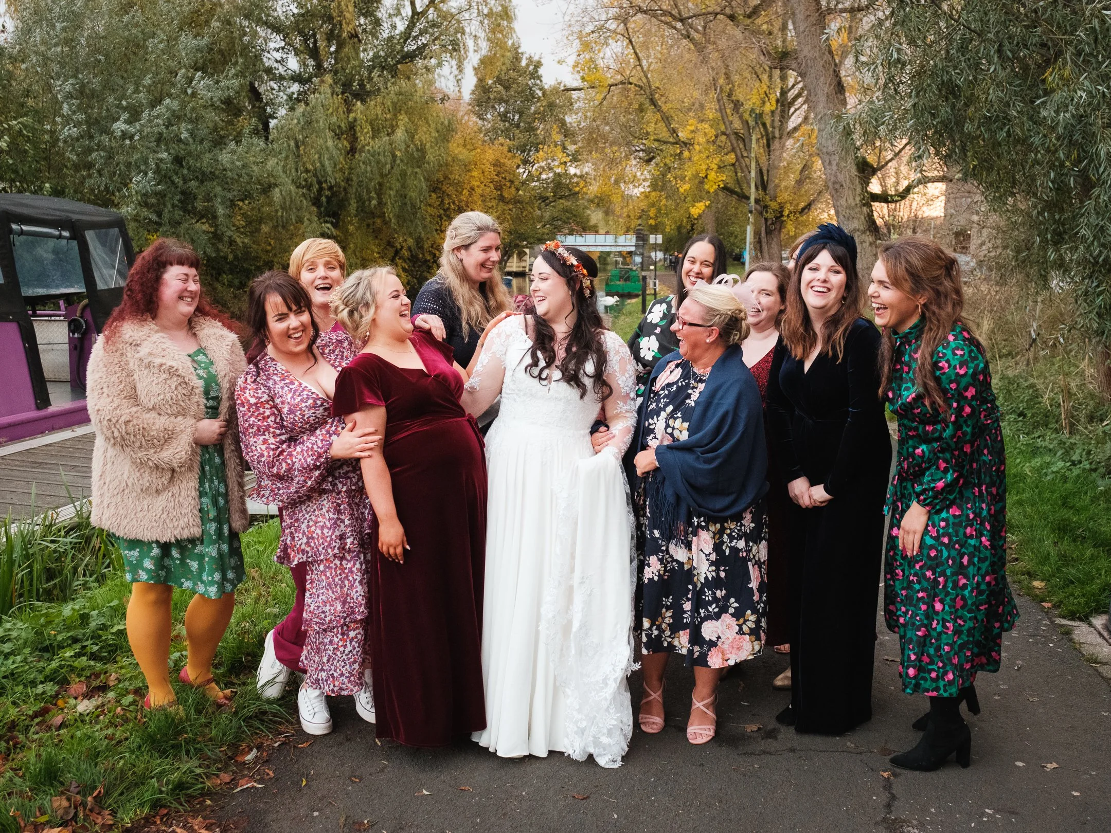 Group of women at a wedding celebration outdoors, smiling and laughing, with autumn trees in the background.