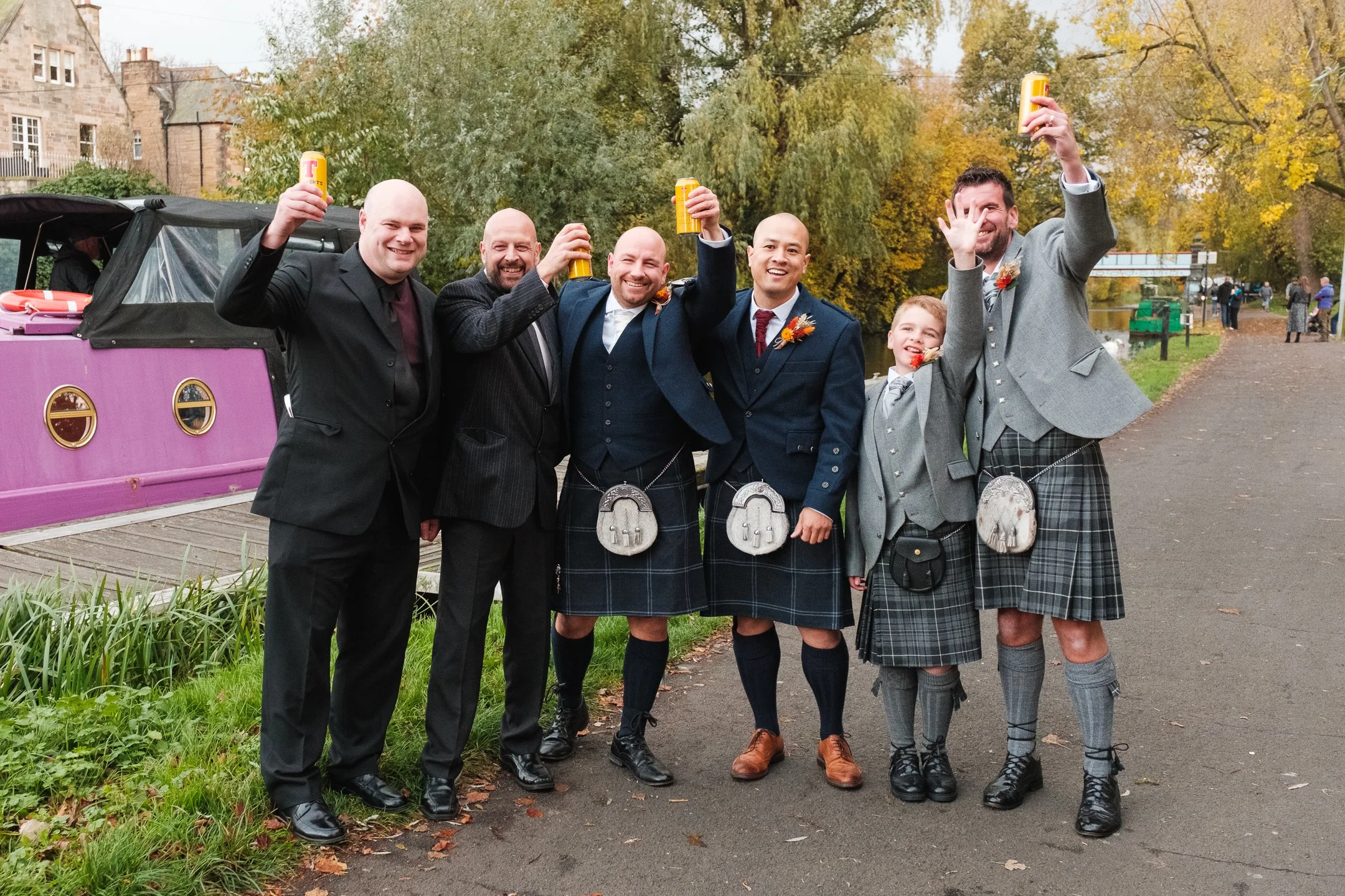 Group of six people, five in kilts and jackets, celebrating outdoors with drinks near a canal or river with trees and historic buildings