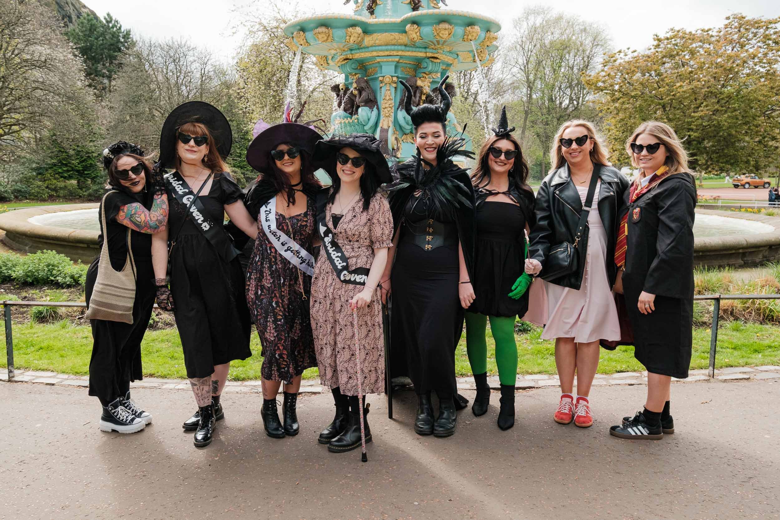 A group of women dressed as witches standing in front of Ross fountain in Princes street gardens in Edinburgh, with trees and a cloudy sky in the background.