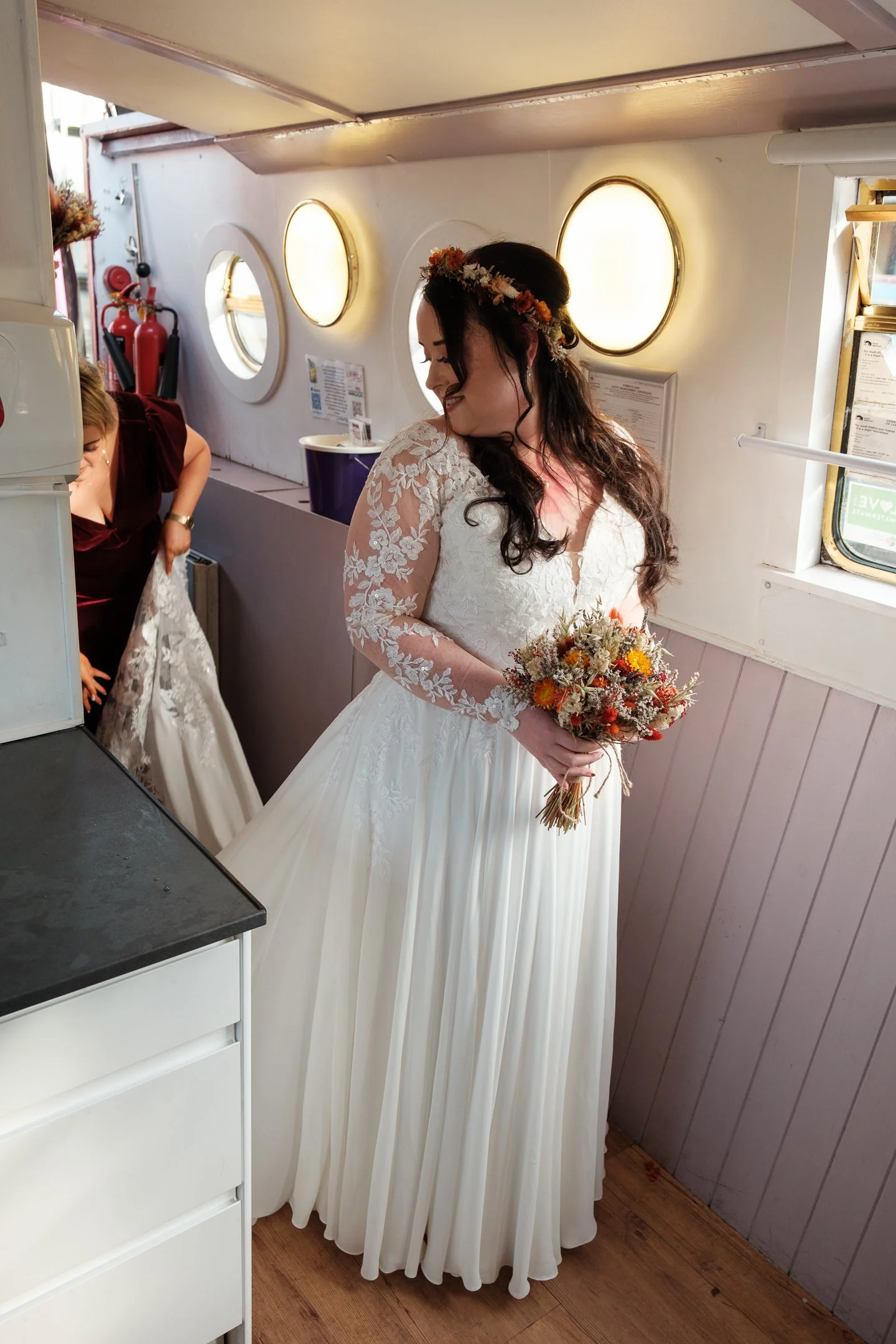 A bride in a white lace wedding dress holding a bouquet, standing inside a small boat or ship in Edinburgh. She has long dark hair, a floral crown, and is smiling while looking down before walking down the aisle.
