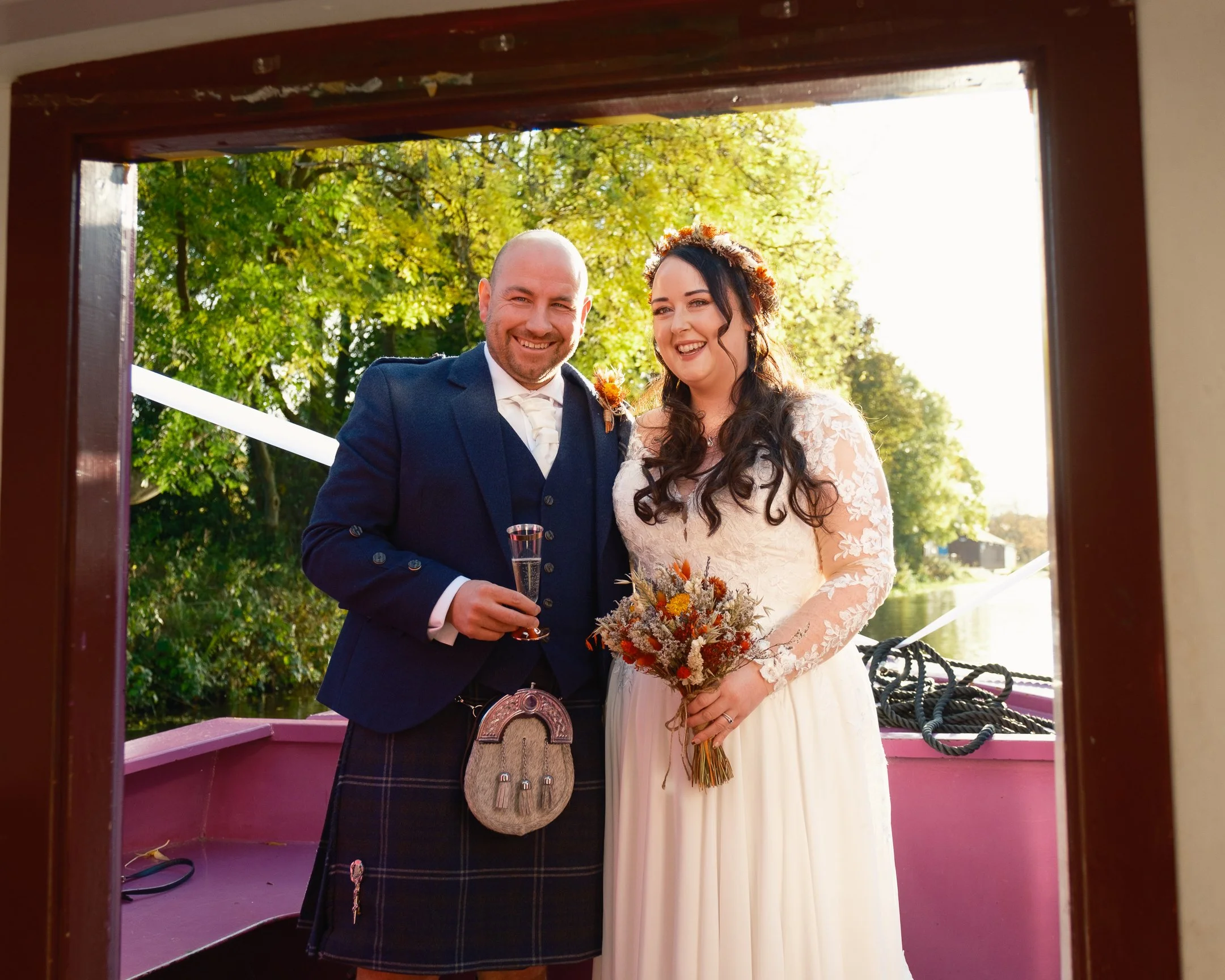 A bride and groom standing on a boat with greenery and water in the background, smiling and dressed in wedding attire, holding drinks and a bouquet.