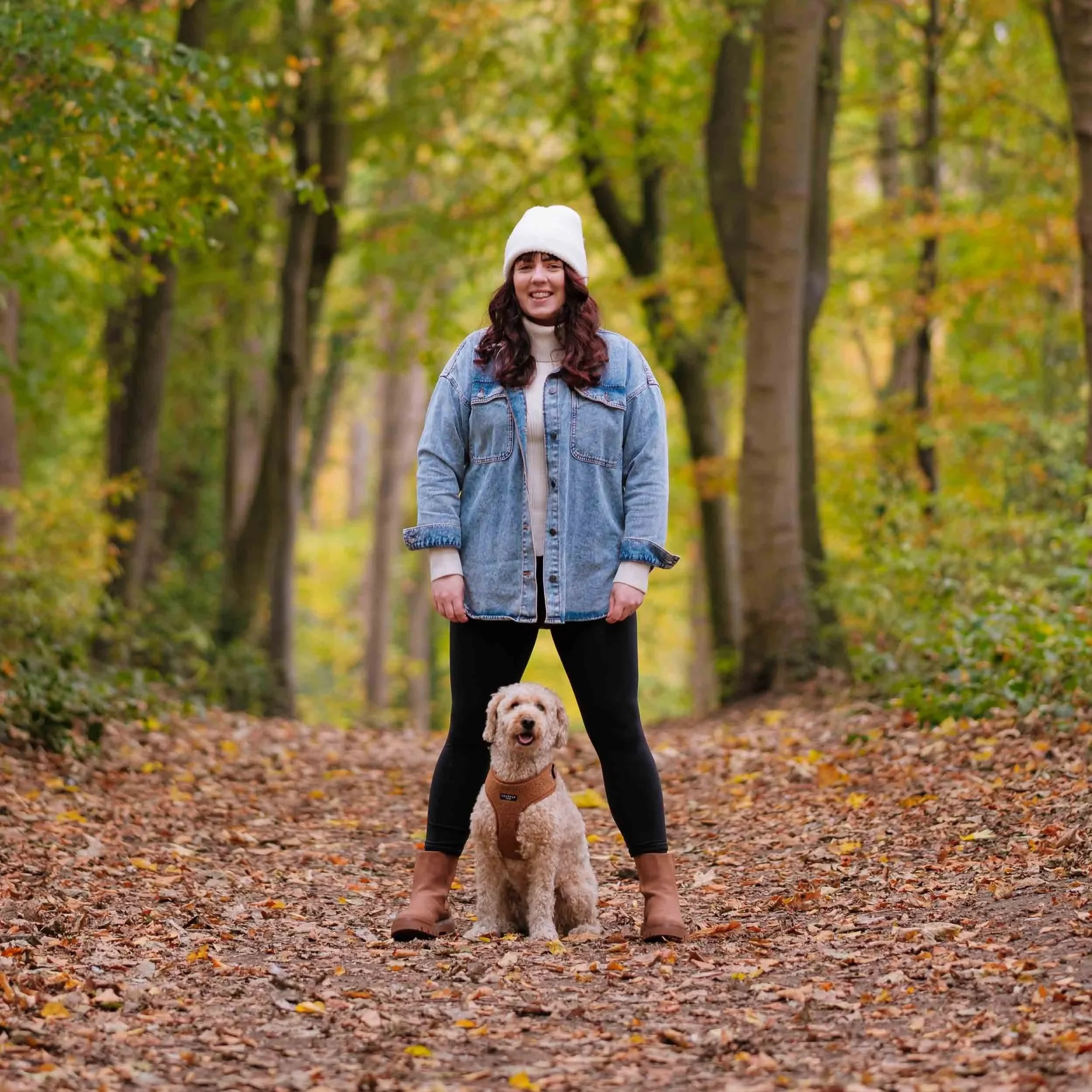 A woman in a white beanie, denim jacket, black leggings, and brown boots standing on a leaf-covered path in Dalkeith country park, with a small cockapoo dog sitting in front of her. The forest has tall trees with green and yellow leaves.