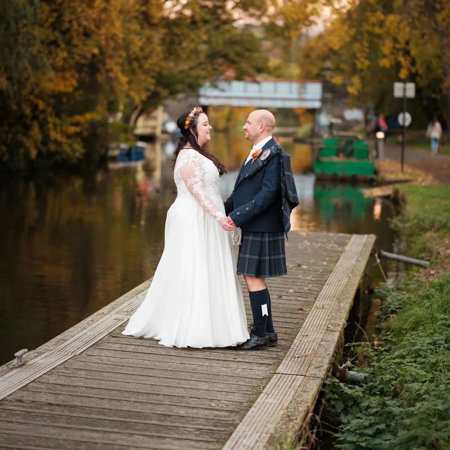 A bride and groom holding hands and gazing at each other on a wooden dock beside a river in Edinburgh with autumn trees and a bridge in the background during their wedding photoshoot.