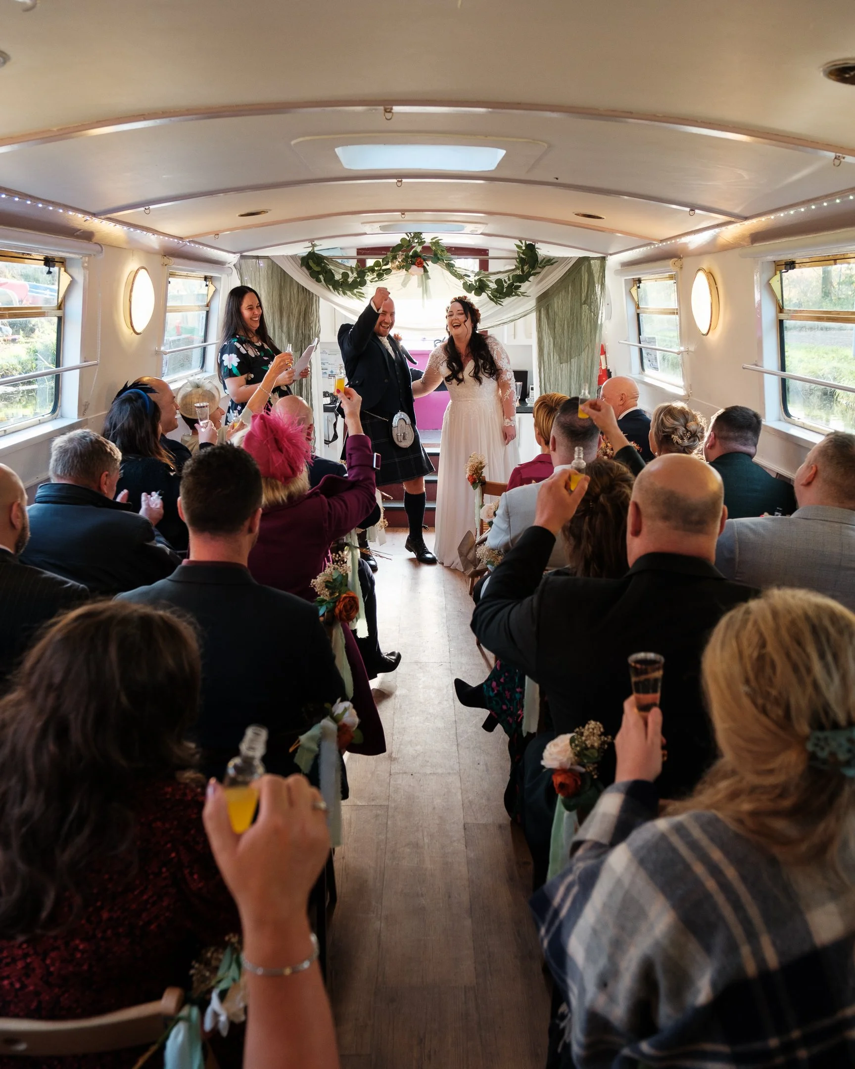 Bride and groom at a wedding ceremony inside a canal boat with seated guests celebrating and raising drinks.