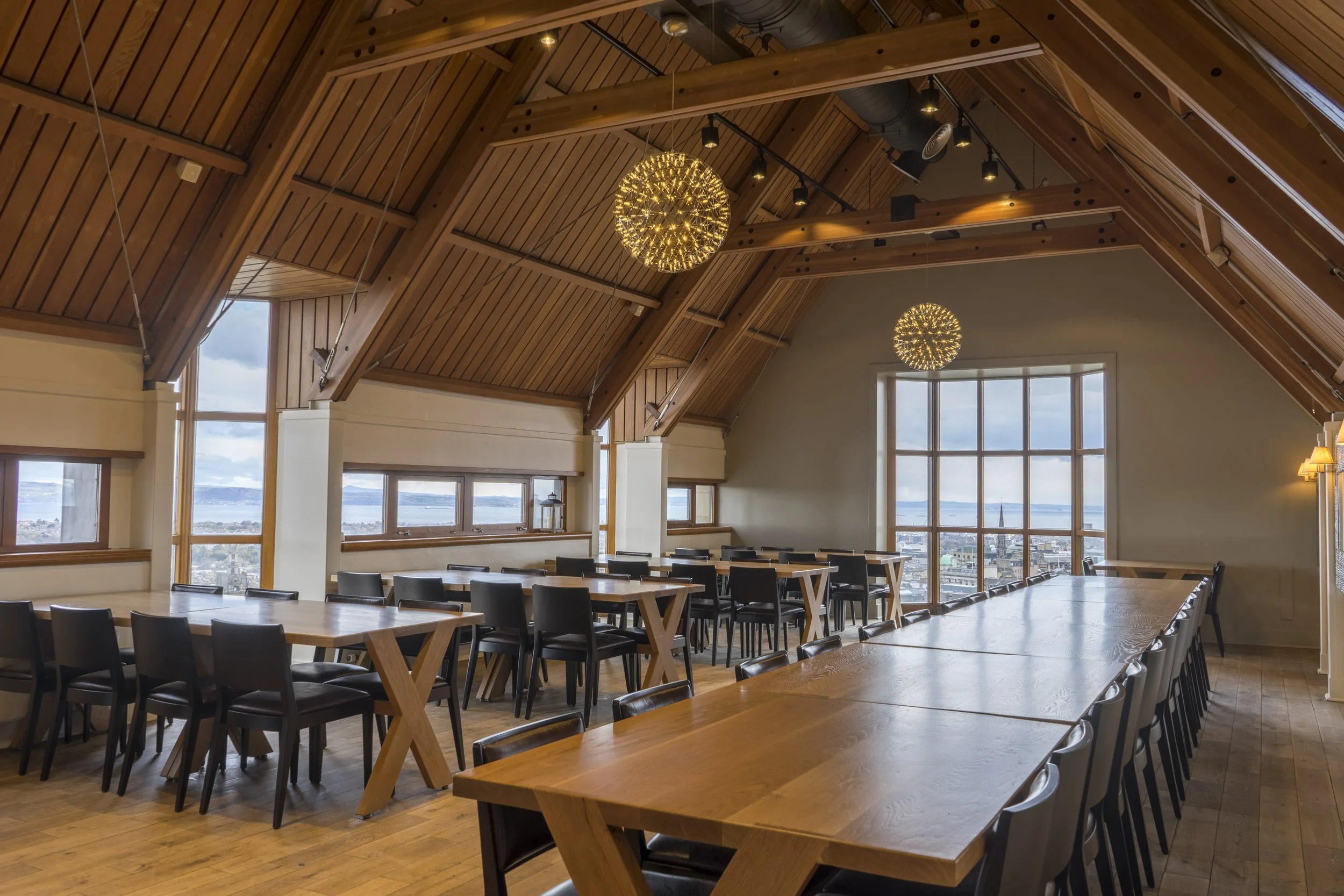 An interior photography of a spacious modern dining and events area in the Jacobite room in Edinburgh Castle with wooden tables and black chairs, large windows providing a view of Edinburgh city, decorative pendant lights hanging from a ceiling.