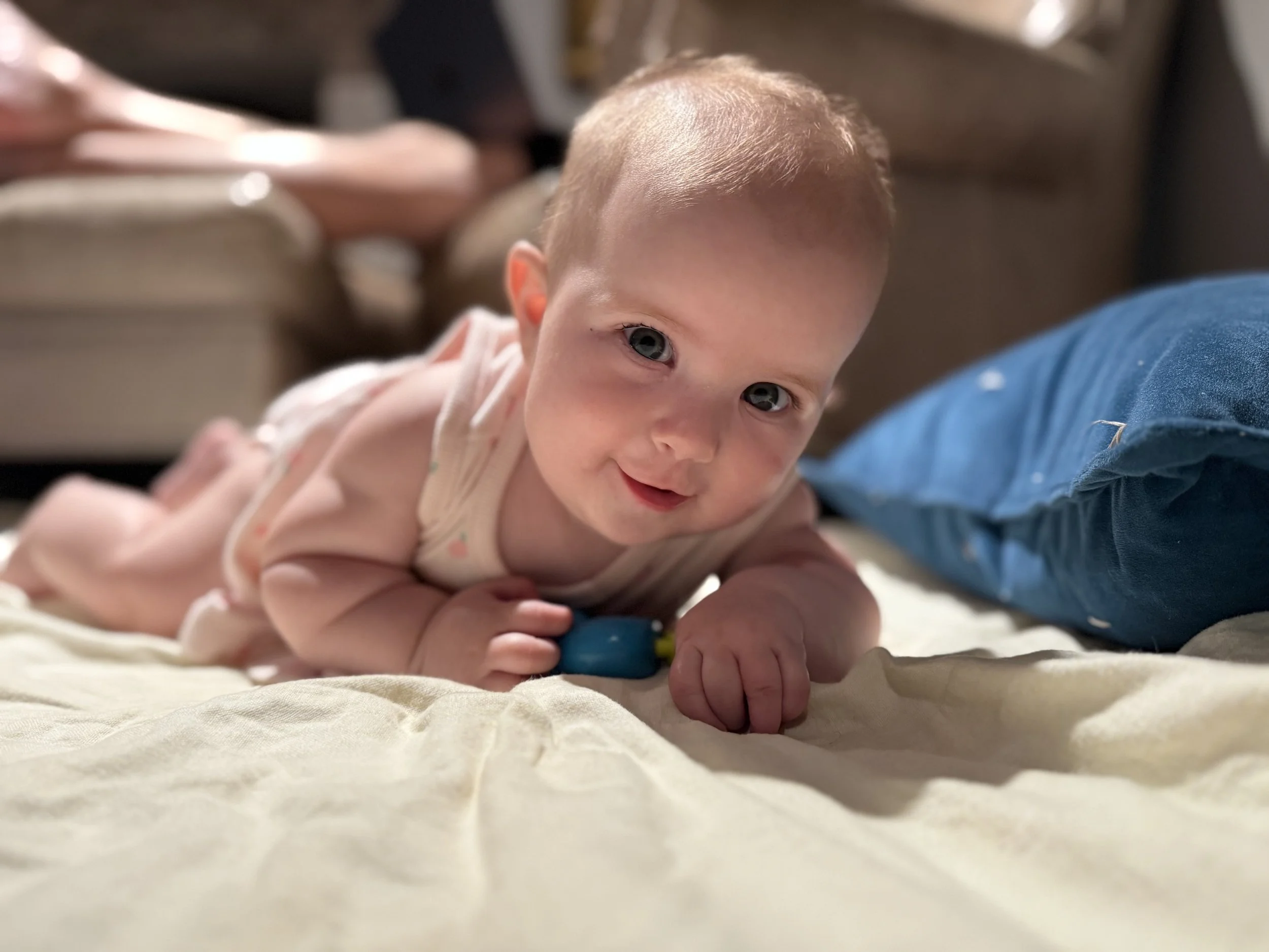 A young baby with light skin and blue eyes lying on a bed, facing the camera with a smile, with a blue pillow nearby and blurred background.