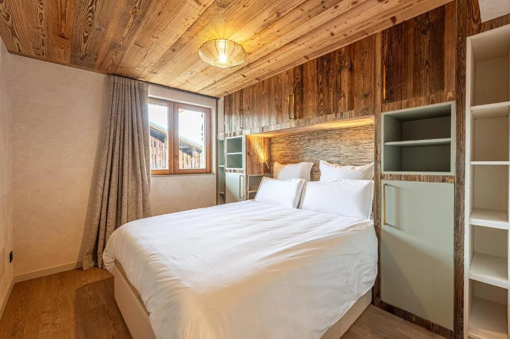 Chambre avec un lit double blanc, murs en bois, fenêtre avec des rideaux, éclairage tamisé, décor chaleureux.