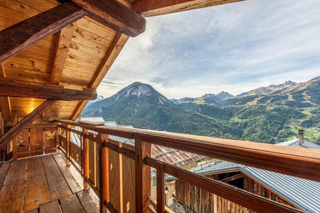 Balcon en bois d'une cabane en montagne avec vue sur des montagnes verdoyantes et un sommet rocheux.