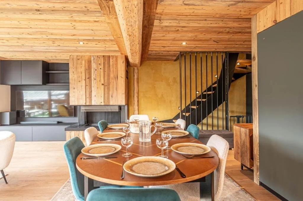 Salle à manger avec une table ronde en bois, assiettes, verres et carafes, autour de chaises de différentes couleurs. Cuisine moderne en arrière-plan, escalier noir en métal, murs en bois et en jaune, plafond en bois.