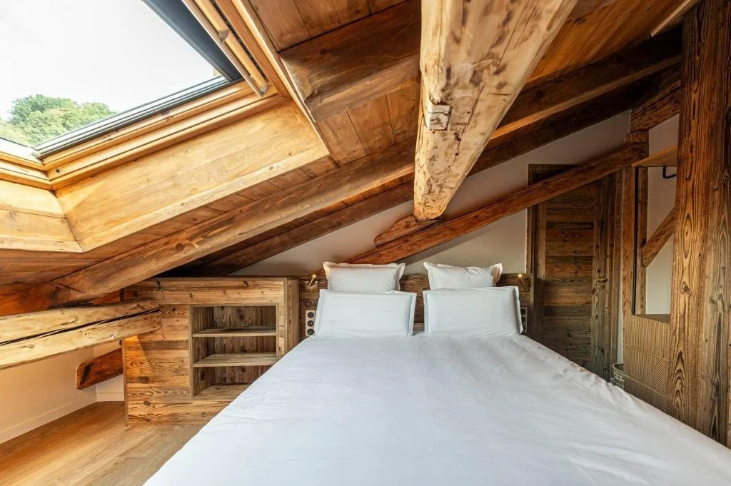 Chambre avec toit en bois et poutres apparentes, lit avec draps blancs, fenêtre de toit laissant entrer la lumière naturelle, murs et meubles en bois rustic.