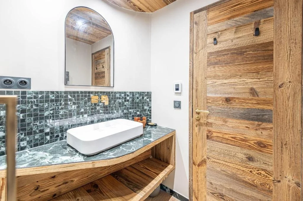 Petite salle de bains avec un plan de travail en marbre vert, un évier carré blanc, un miroir arqué, un mur en mosaïque sombre, une porte en bois, des interrupteurs modernes et des petits accessoires de salle de bains.