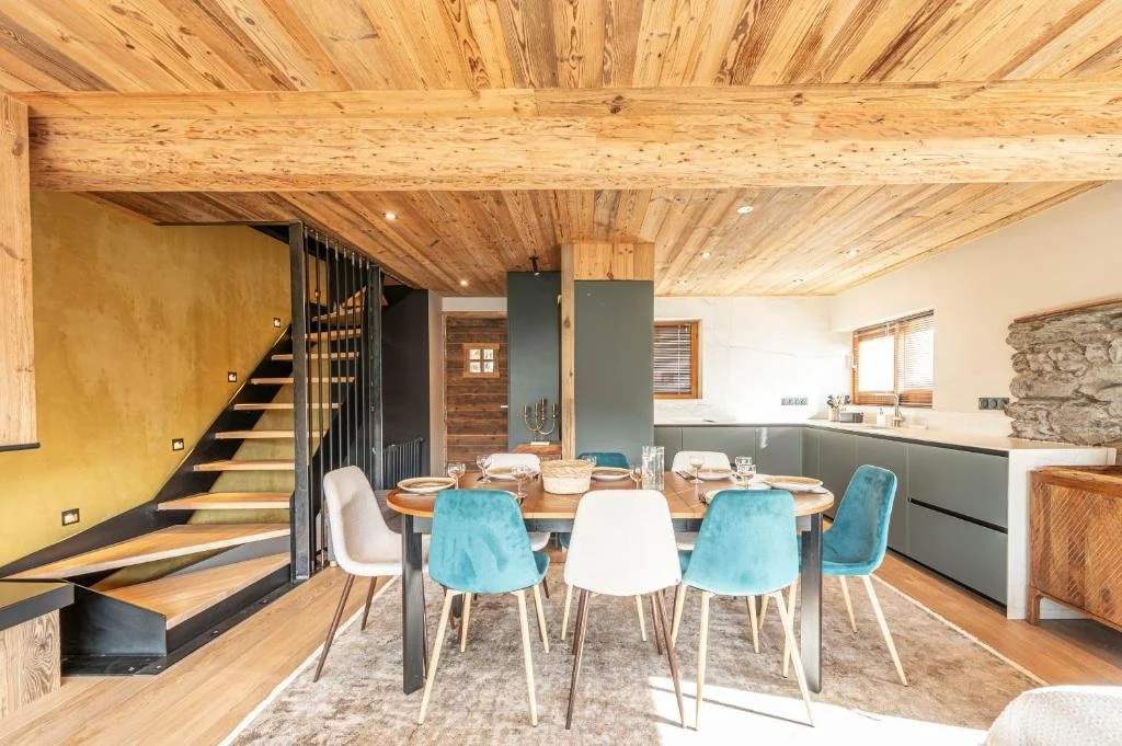 Une salle à manger dans une maison en bois avec un plafond en bois, un ensemble de chaises bleues et blanches autour d'une table, des fenêtres avec des volets en bois, et une cuisine en arrière-plan.