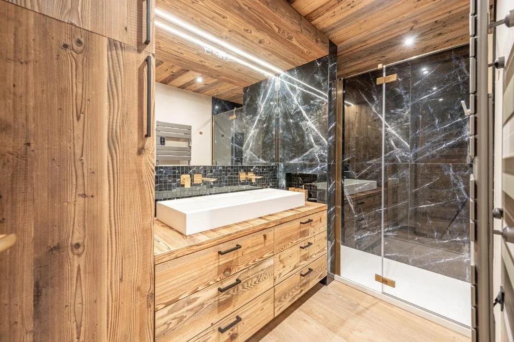 Une salle de bain élégante avec un lavabo rectangulaire blanc sur un meuble en bois, des murs en marbre noir avec veines blanches, une cabine de douche en verre avec porte à verrou, et un plafond en bois avec des spots lumineux encastrés.