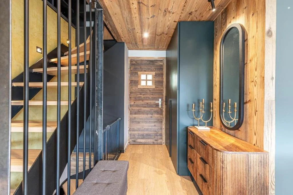 Entrée d'une maison avec un escalier en bois et métal, une porte en bois, un meuble en bois avec un miroir et deux candélabres en or.
