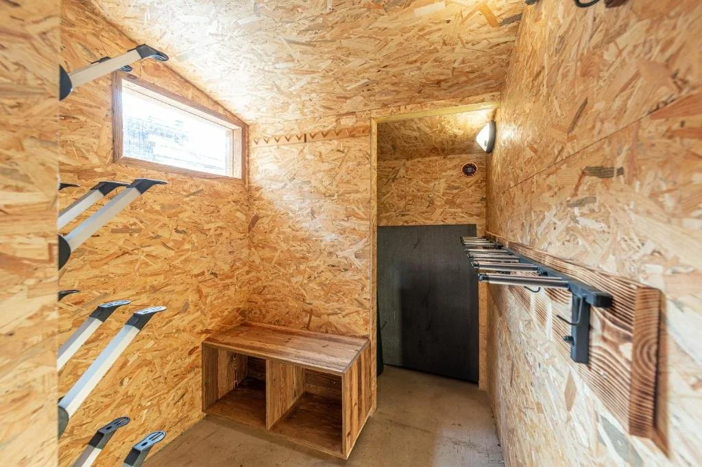 Petit atelier ou débarras avec parois en panneaux de bois, étagères métalliques à gauche, un petit meuble en bois en face, une porte de stockage en arrière, et un éclairage au plafond.