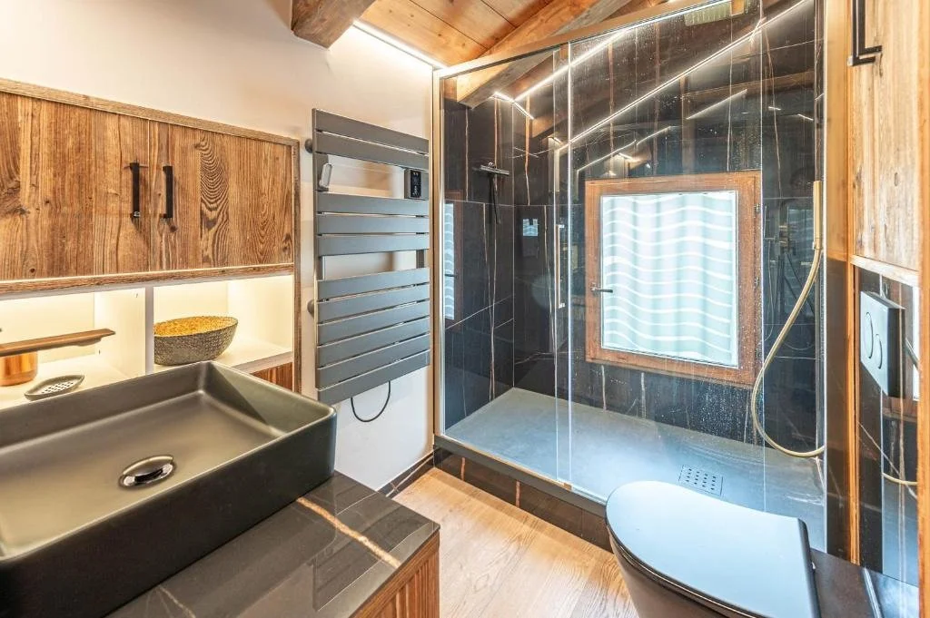 Salle de bains moderne avec douche à l'italienne en verre, lavabo noir dans un meuble en bois, toilettes noires, mur en carreaux noirs avec une grande fenêtre en bois, lumière LED.