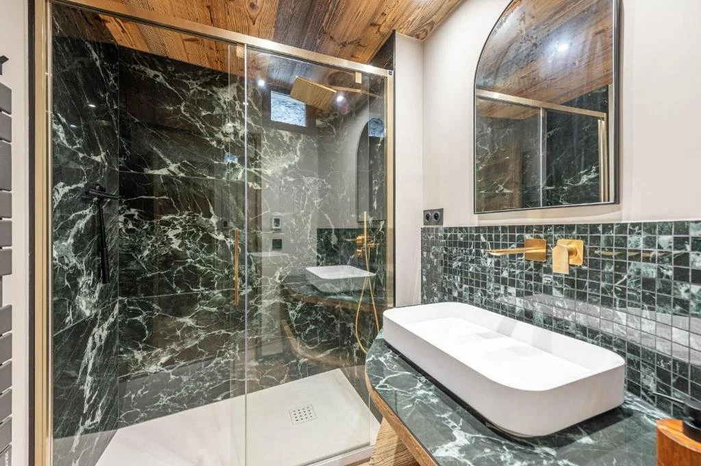 Salle de bain avec mur de douche en marbre noir à veines blanches, cabine de douche en verre, lavabo rectangle blanc sur comptoir en marbre vert, miroir mural grand en forme d'arche, robinetterie dorée, plafond en bois, carrelage mosaïque vert foncé 