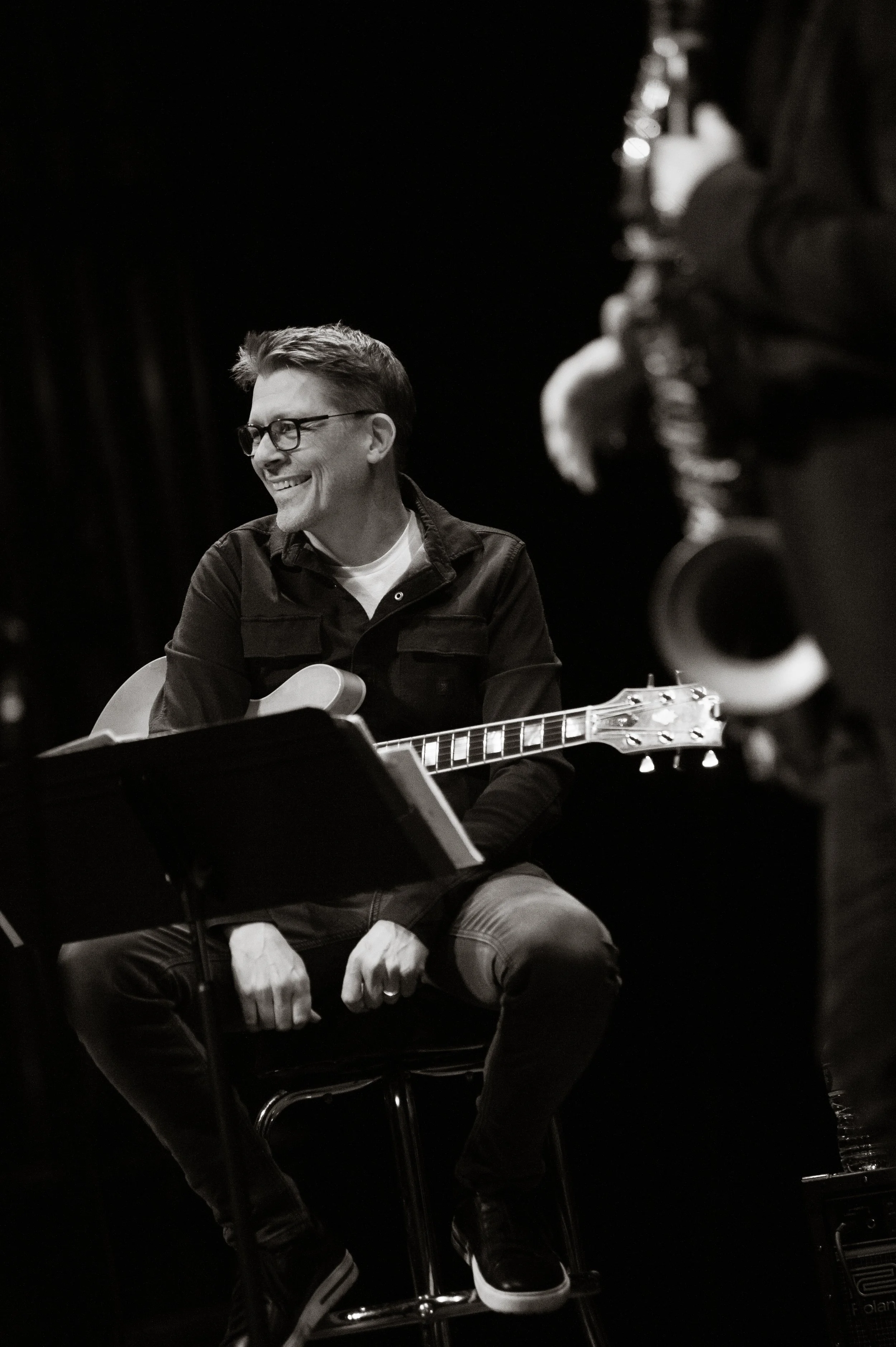 Musician Scott Hesse playing an acoustic guitar on stage, smiling, wearing glasses and a black jacket, in a black-and-white photo.