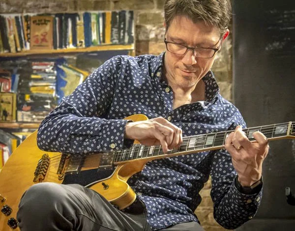 Musician Scott Hesse sitting and playing an electric guitar in a room with bookshelves and brick wall.