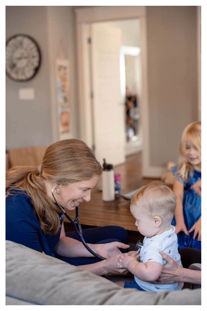 A female healthcare professional, possibly a nurse or pediatrician, using a stethoscope to listen to a young child's chest, with two young children watching in a cozy, home setting.