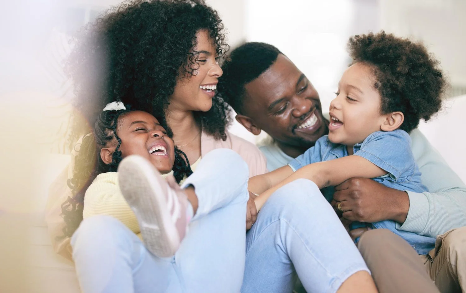 Family of four, two parents and two young children, laughing and playing together on a sofa, enjoying a happy moment at home.